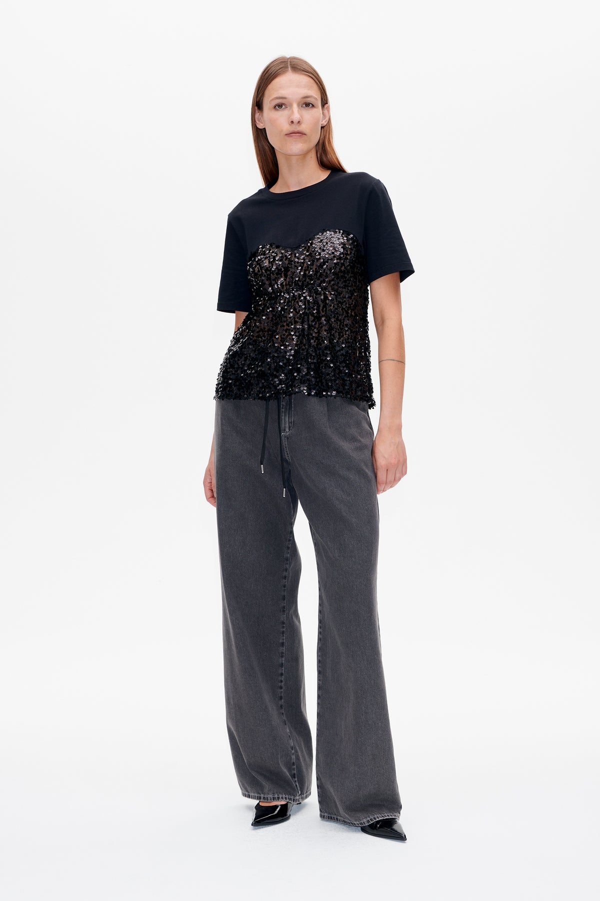 Front view of a model wearing a black short-sleeved T-shirt with a dense sequin panel and charcoal wide-leg jeans.