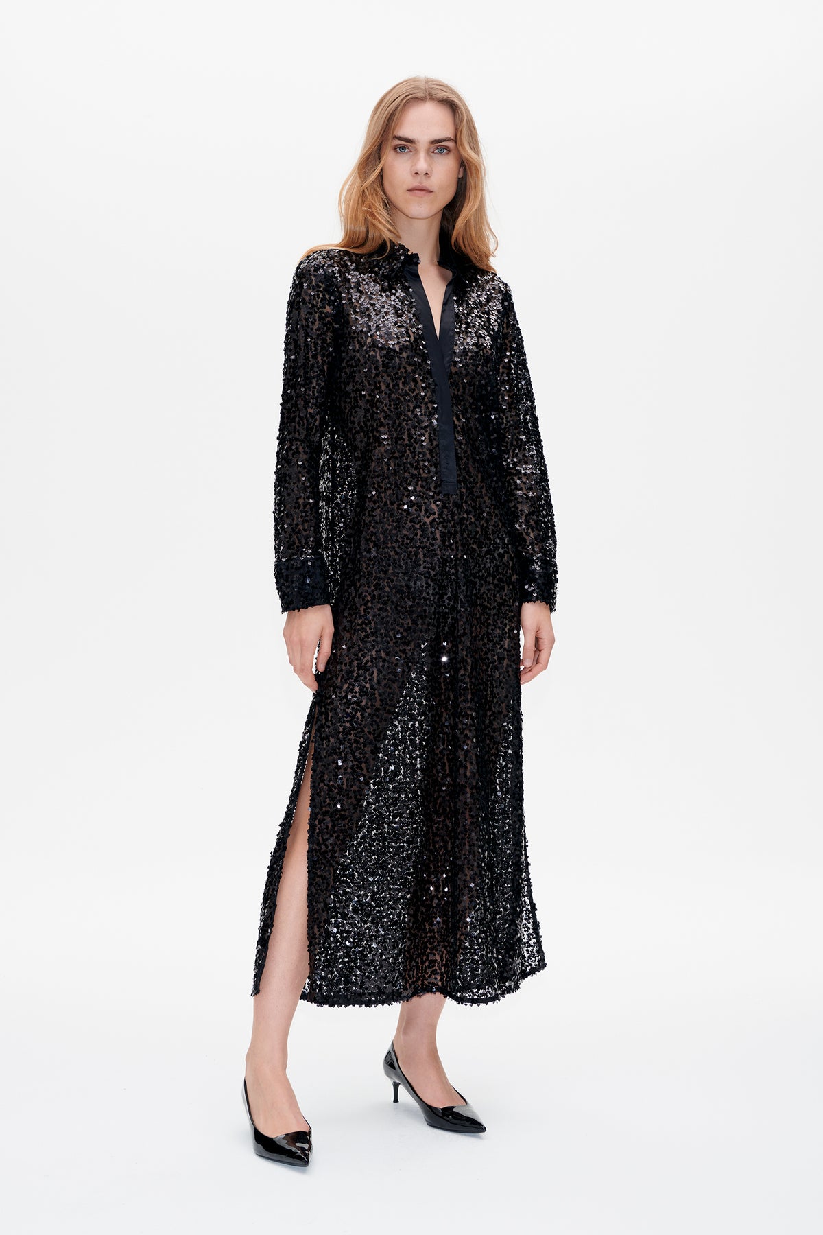 Model wearing a black sheer sequin-embellished long-sleeved dress with a collared V-neck and high side slit, shown from the front.
