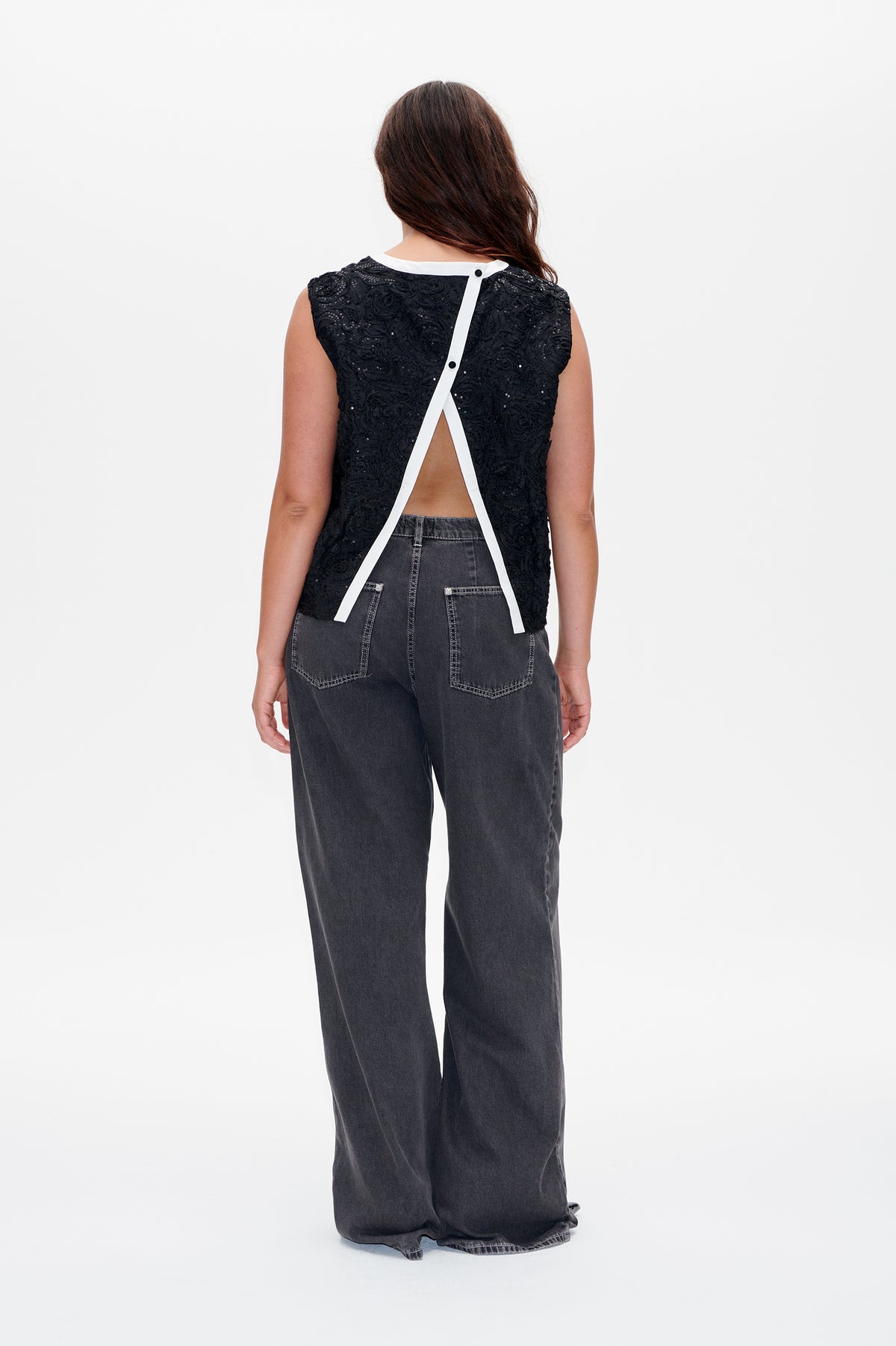 Back view of a black sleeveless top with textured floral embellishment, white crisscross back trim and an open panel, worn with charcoal wide-leg jeans.