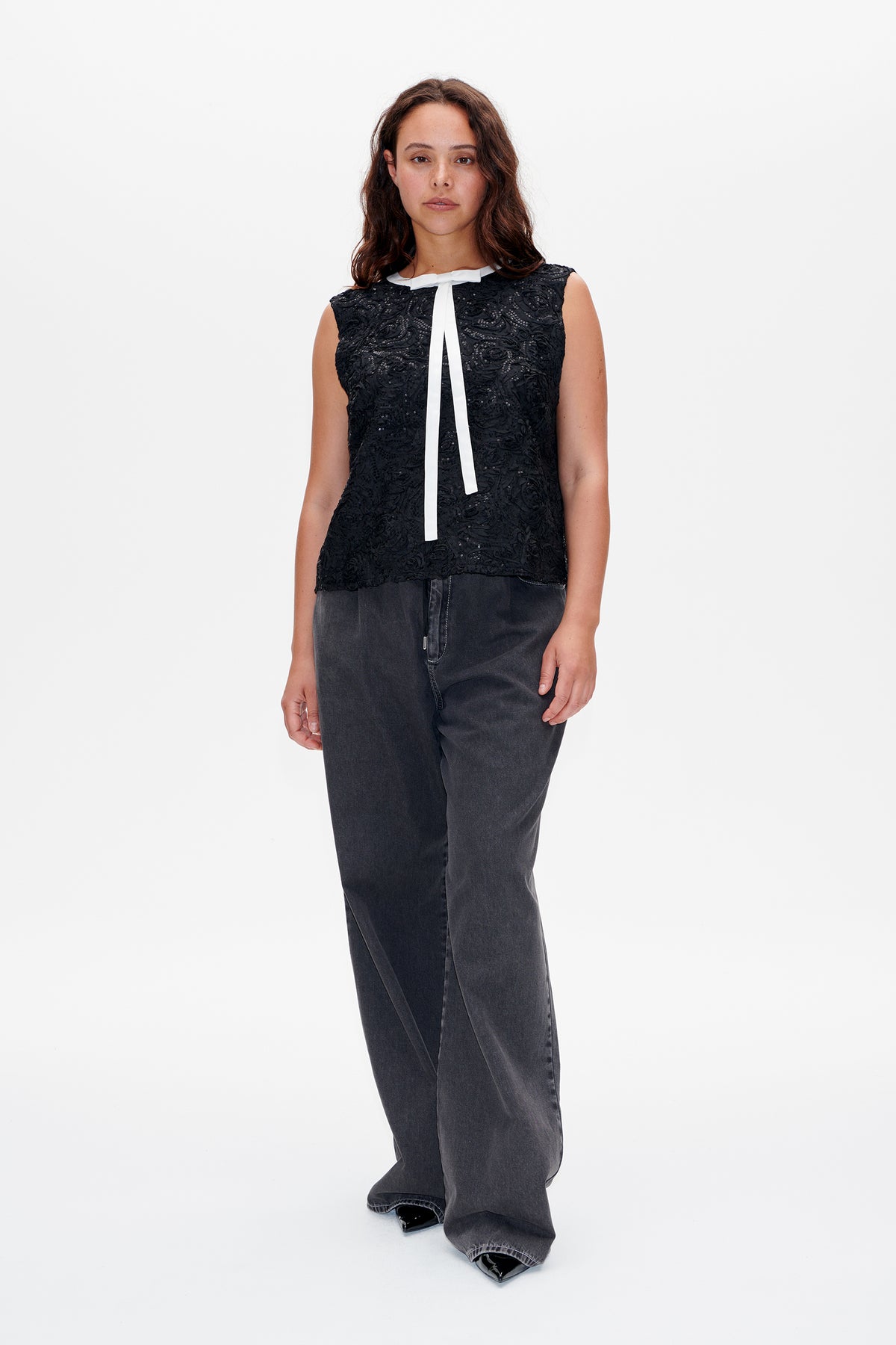Front view of a model wearing a black sleeveless top with textured floral embellishment, a white collar and long ribbon ties, and charcoal wide-leg jeans.