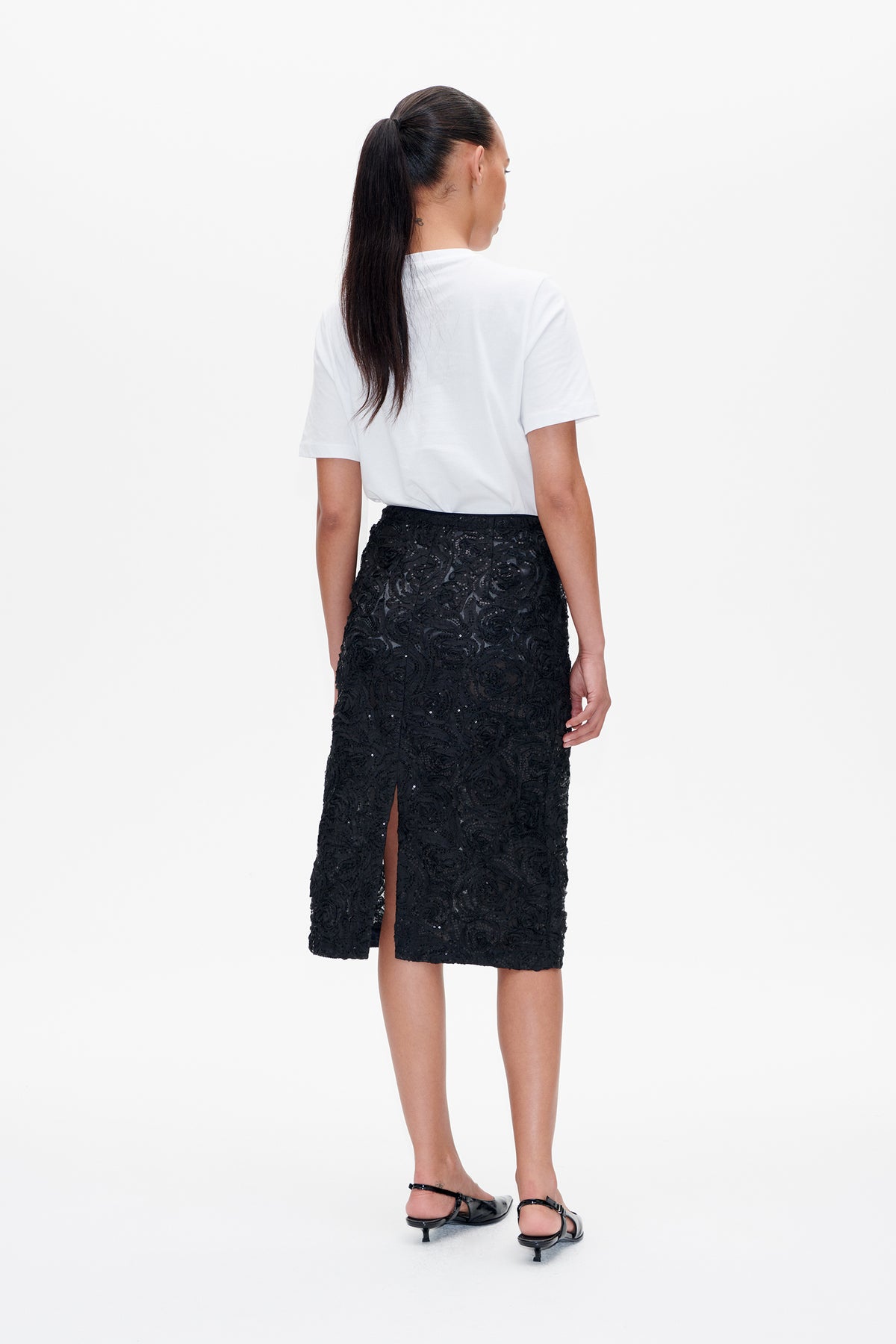 Back view of a model wearing a white short-sleeved T-shirt and a black embellished midi skirt with a slit.