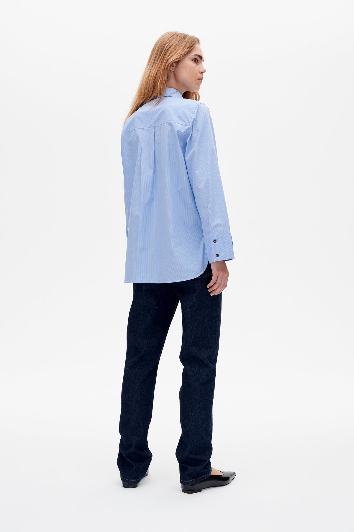 Modell trägt eine hellblaue, fein gestreifte Bluse in Dreiviertelansicht von hinten, mit Rückenpasse und Falte, kombiniert mit dunklen Jeans.
