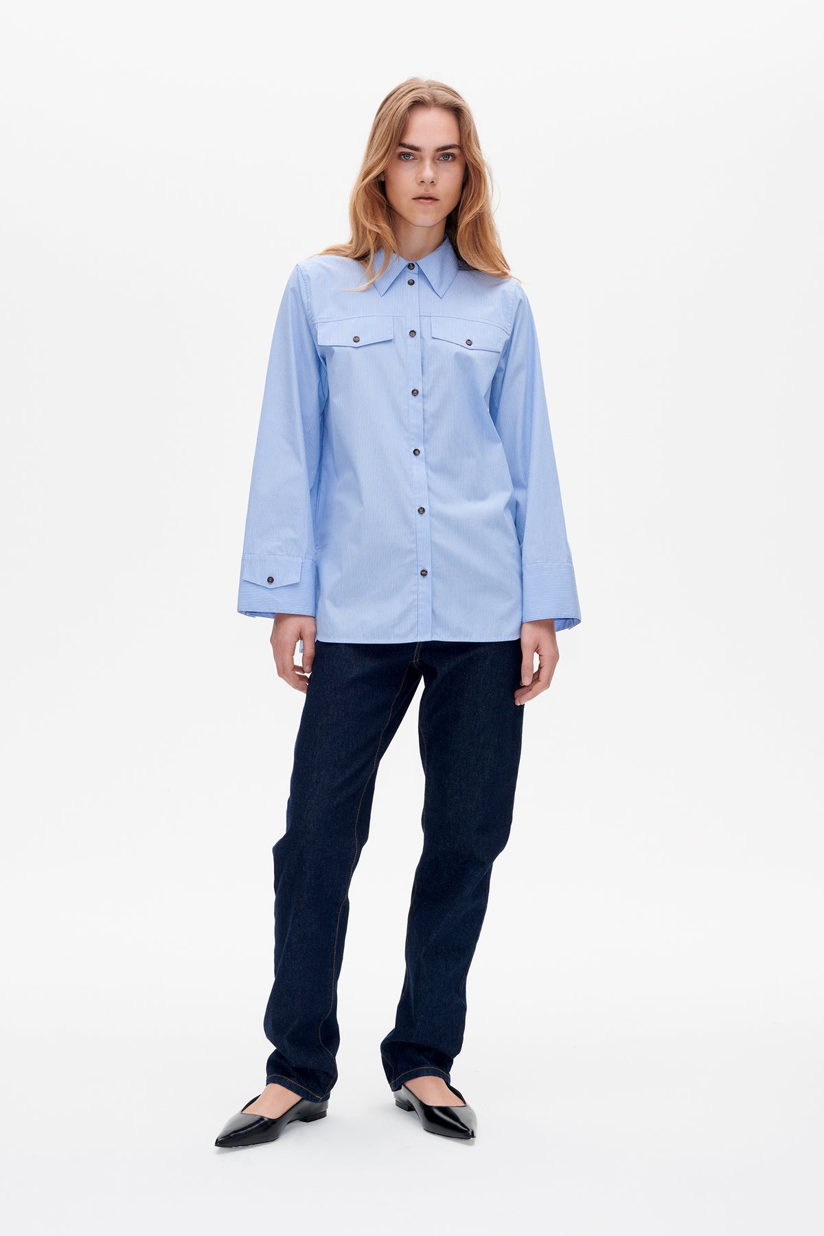 Vorderansicht eines Models in einem hellblauen, fein gestreiften Hemd mit Knopfleiste, Brusttaschen und weiten Manschetten, dunklen Jeans und schwarzen Ballerinas.