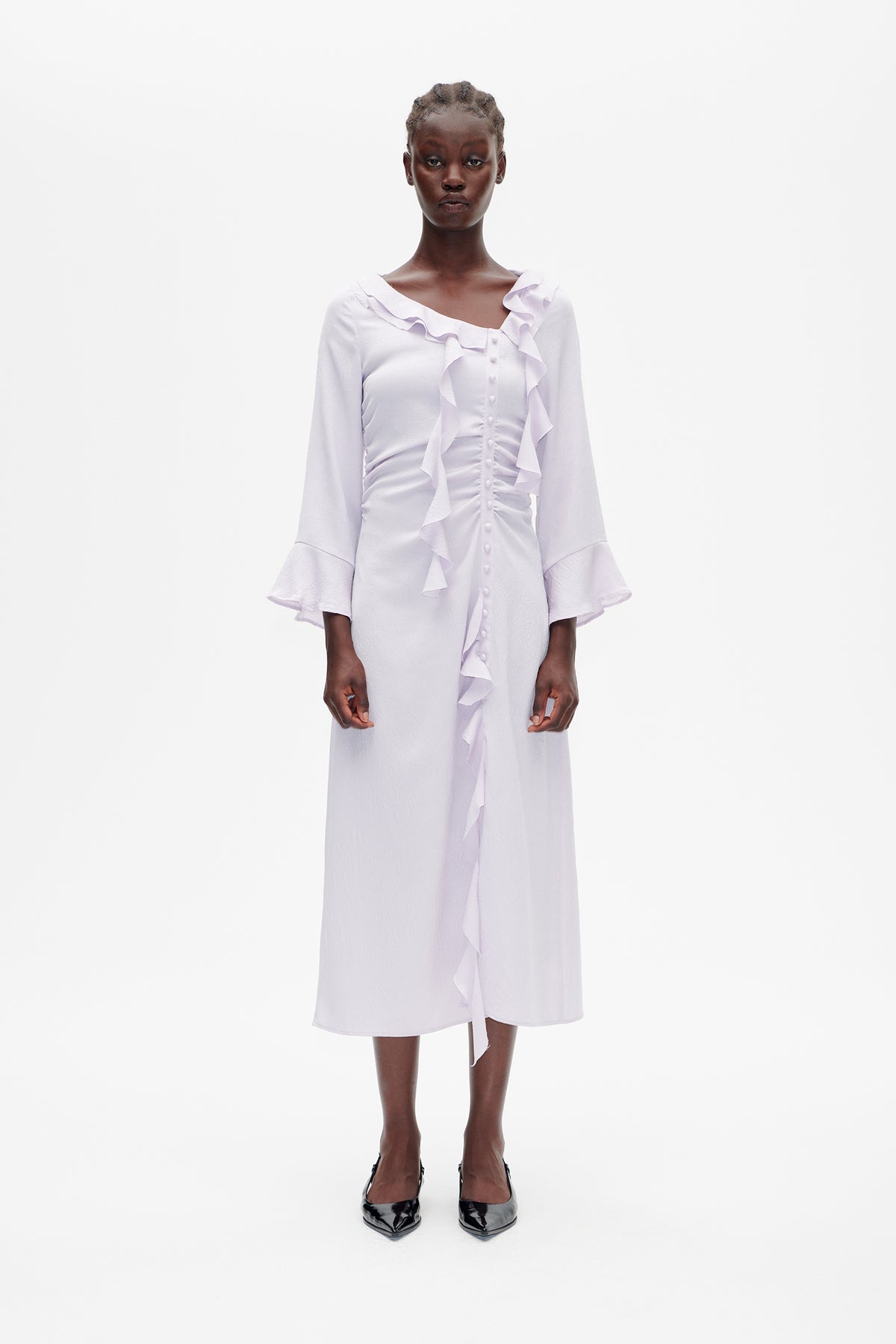 Model wearing a pale lilac midi dress with a ruffled neckline and front, covered buttons, and flared three-quarter sleeves.