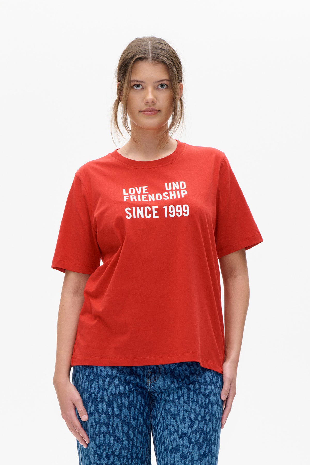 Front view of a model wearing a red short-sleeved T-shirt printed “Love und Friendship Since 1999” with blue patterned jeans.