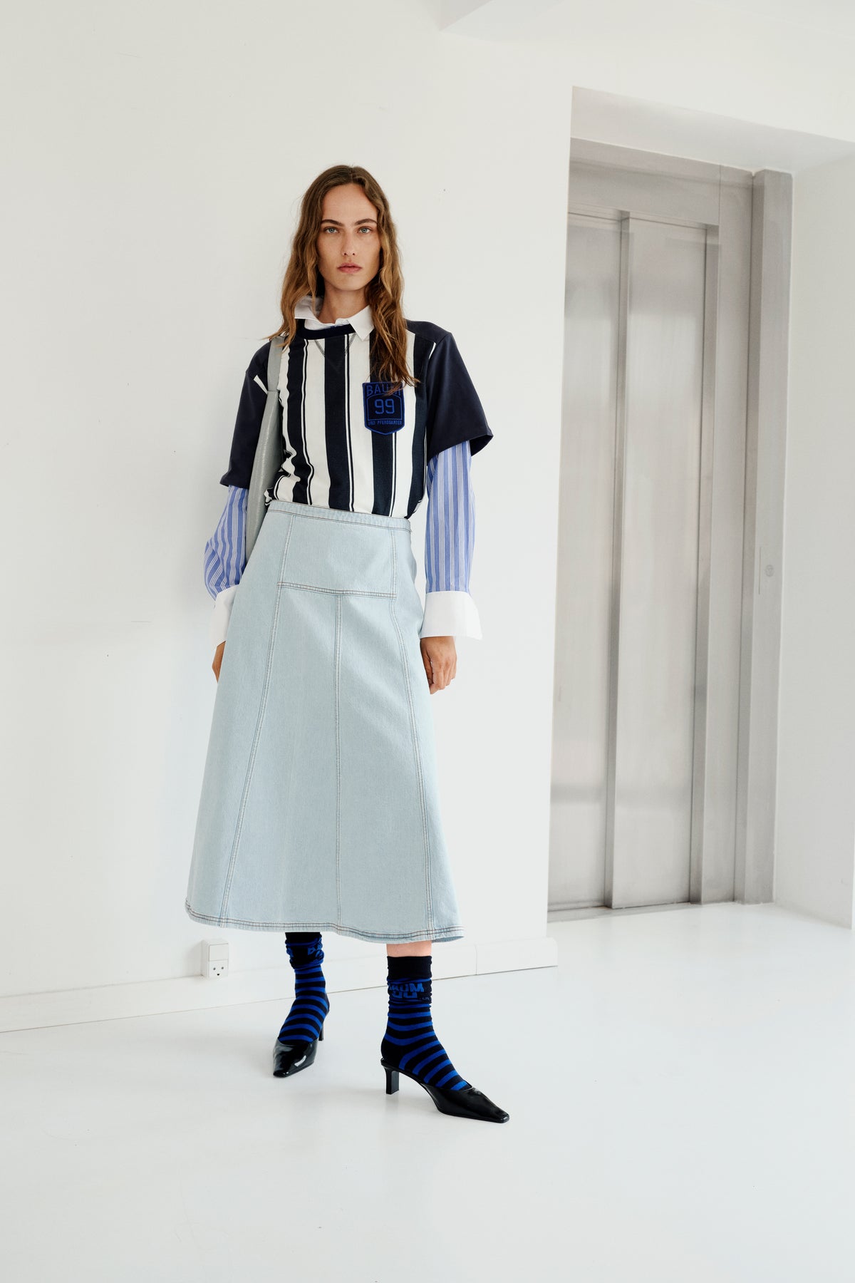 Mannequin dans un intérieur lumineux et minimaliste, portant un haut à manches courtes rayé bleu marine et blanc par-dessus une chemise rayée bleue, une jupe en jean clair et des chaussettes rayées.