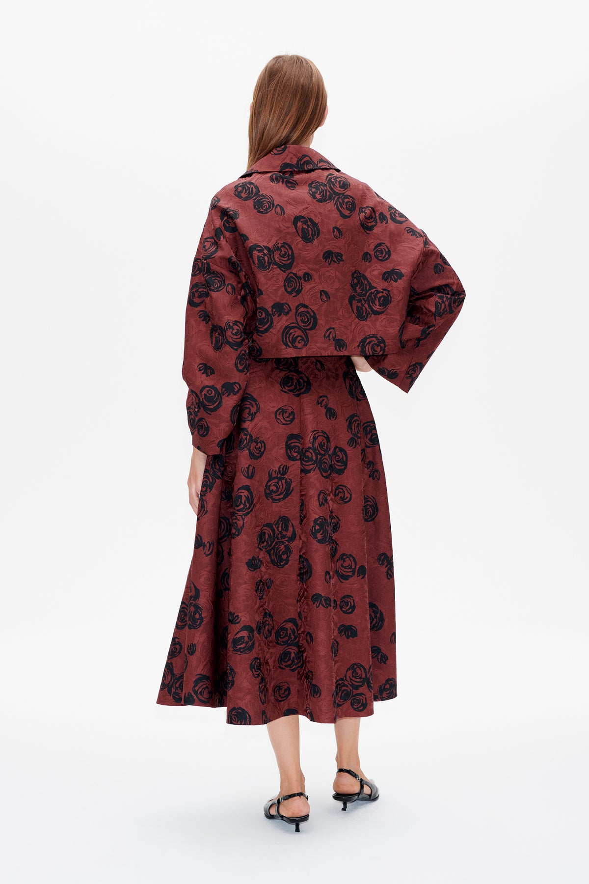 Back view of a model wearing a rust-red, rose-patterned midi dress with a wide collar and loose sleeves.