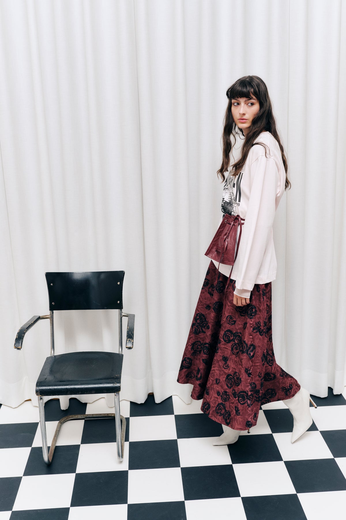 A model wears a white graphic sweatshirt, burgundy rose-print maxi skirt and matching tied waist pouch beside a black chair.