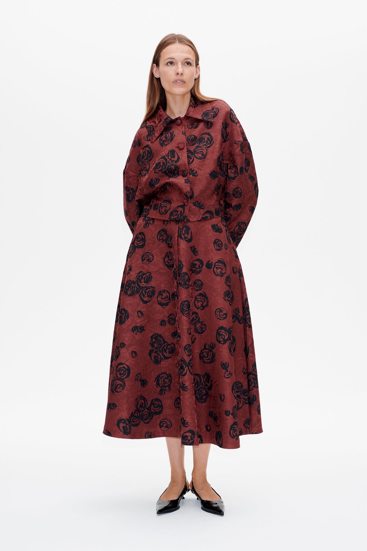 Vorderansicht eines Models, das eine bordeauxrote Jacke mit Knopfleiste und einen passenden Midirock mit schwarzem Rosenprint trägt.