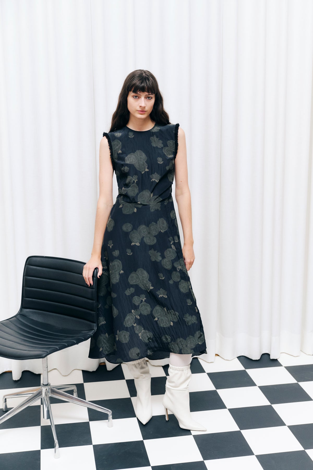 Model wearing a dark sleeveless midi dress with a muted grey floral print and frayed armhole edges, posed beside a chair on a checkerboard floor.