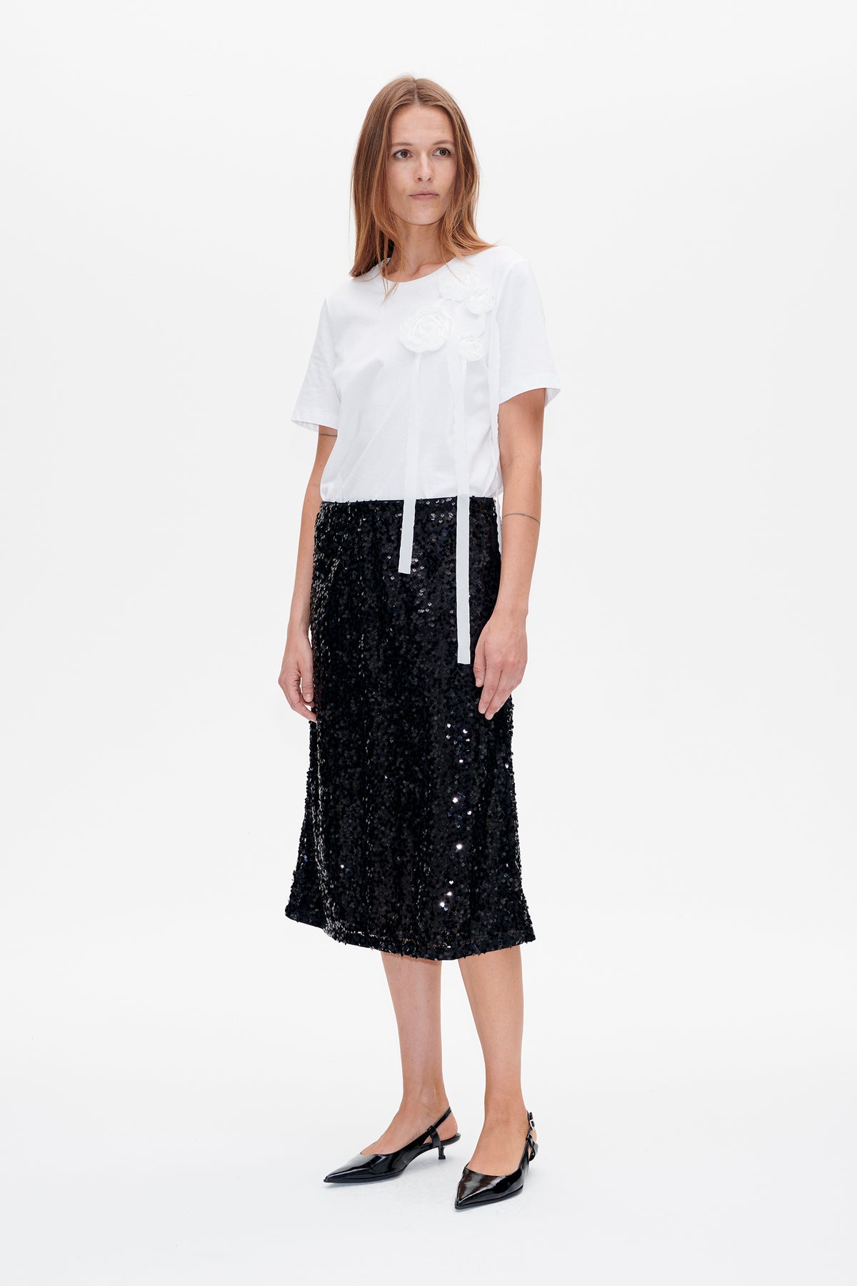 Front view of a white short-sleeved T-shirt with raised floral appliqués and long ribbons, paired with a black sequined midi skirt.