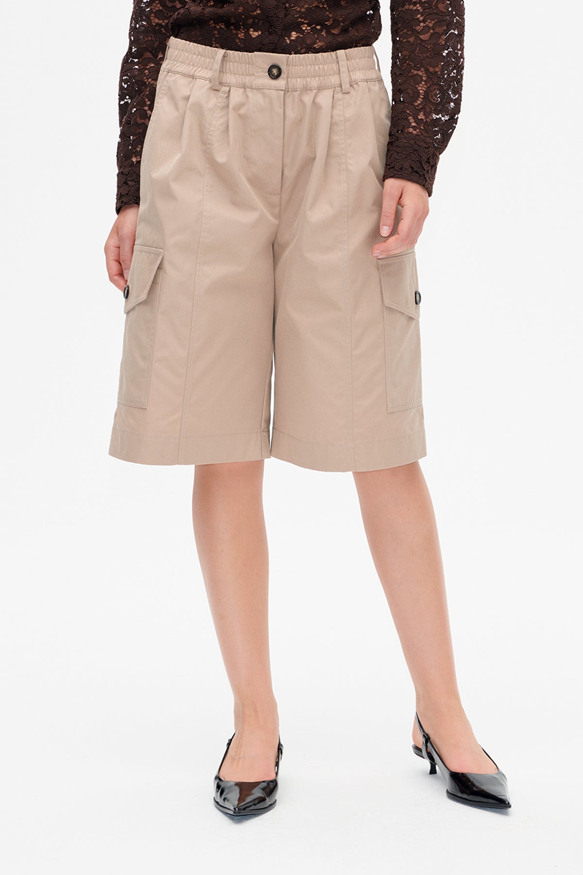Front view of beige knee-length cargo shorts with an elasticated button waistband and large button-fastened side pockets, worn with a dark lace top.