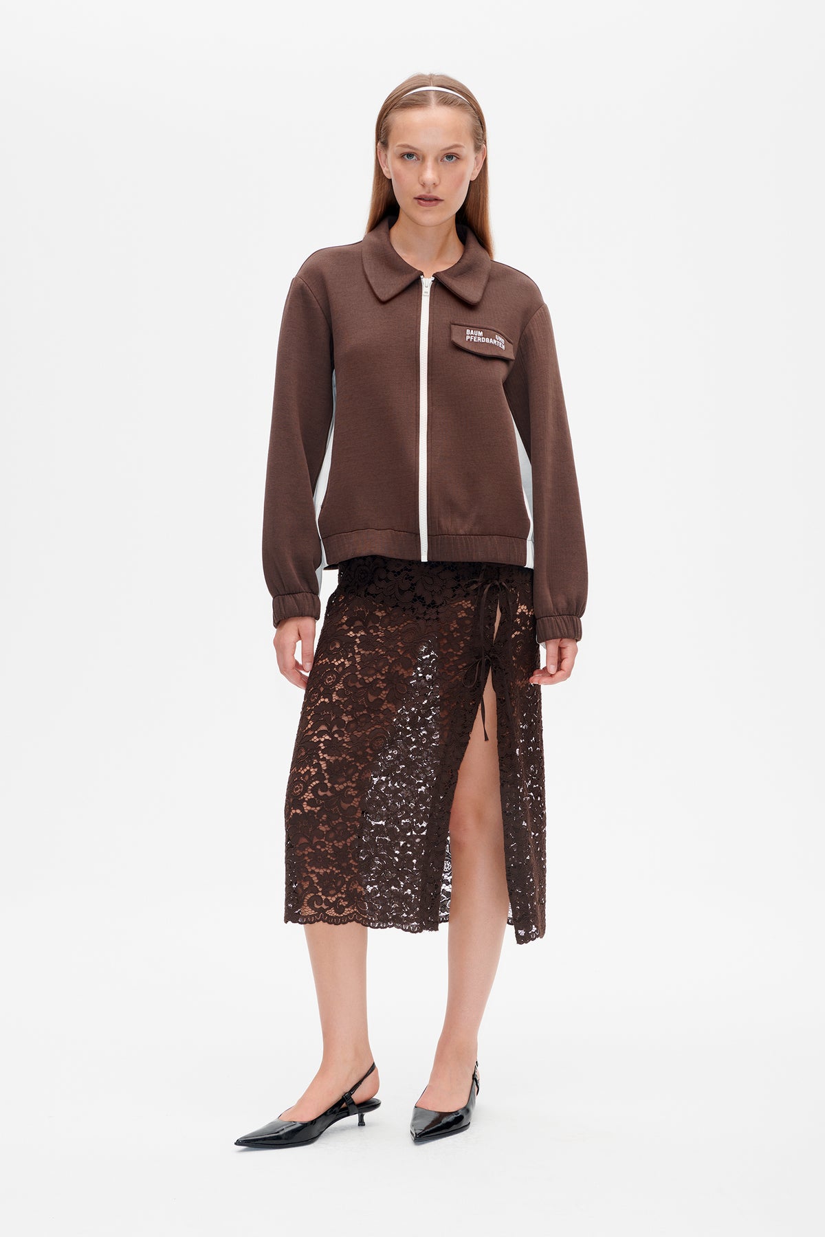 Model wearing a brown zip-front jacket with white trim and chest flap, paired with a dark brown floral lace skirt with side slit.