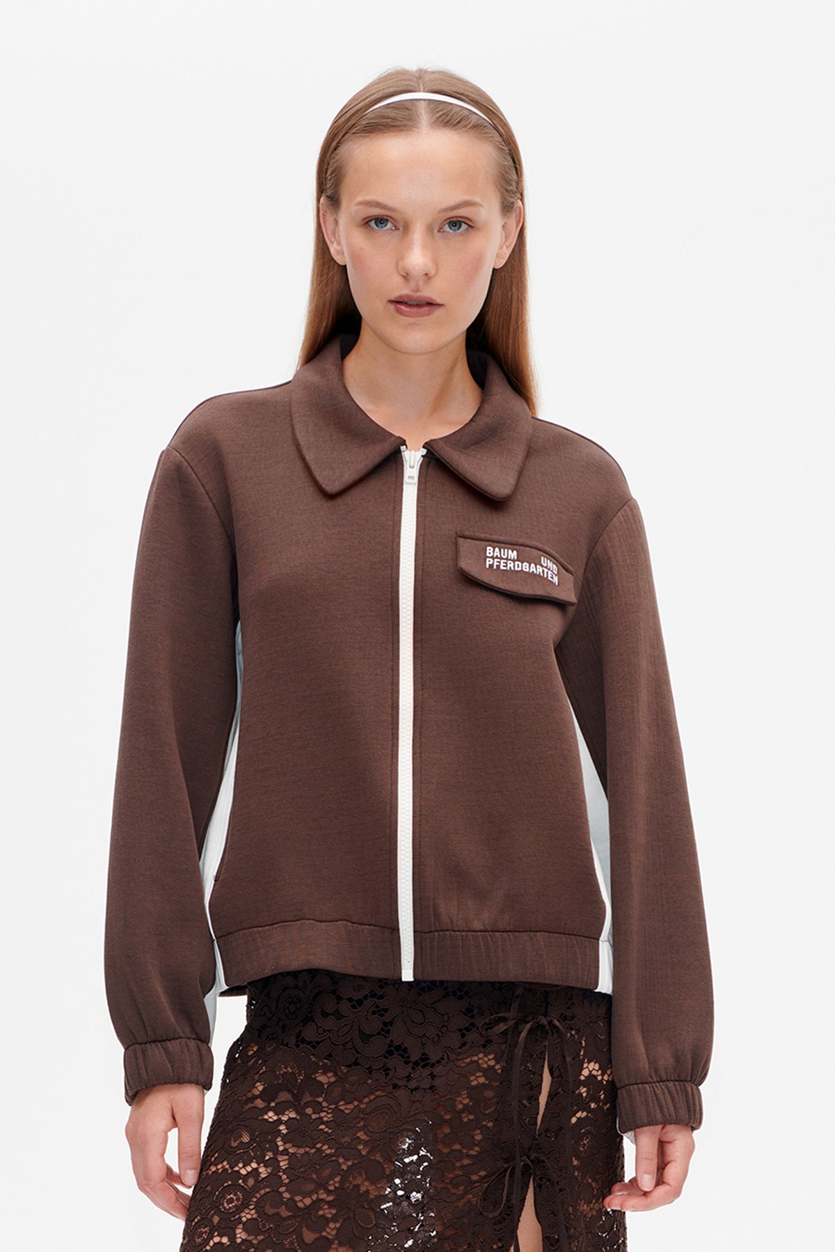 Front view of a brown zip-front jacket with a wide collar, white zipper and side panels, and flap chest pocket, over a dark lace skirt.