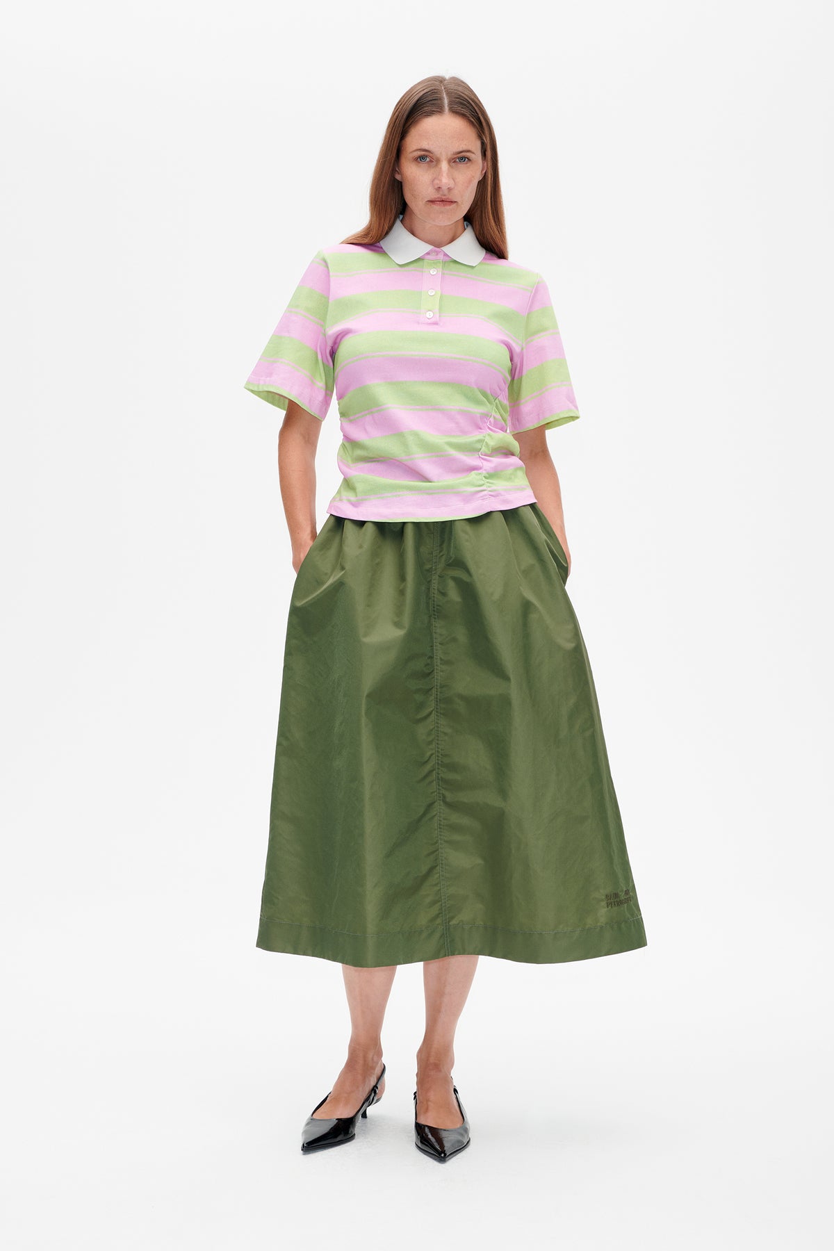 Frontalansicht eines Models in einem rosa- und hellgrün gestreiften Poloshirt mit weißem Kragen und einem olivgrünen Rock.