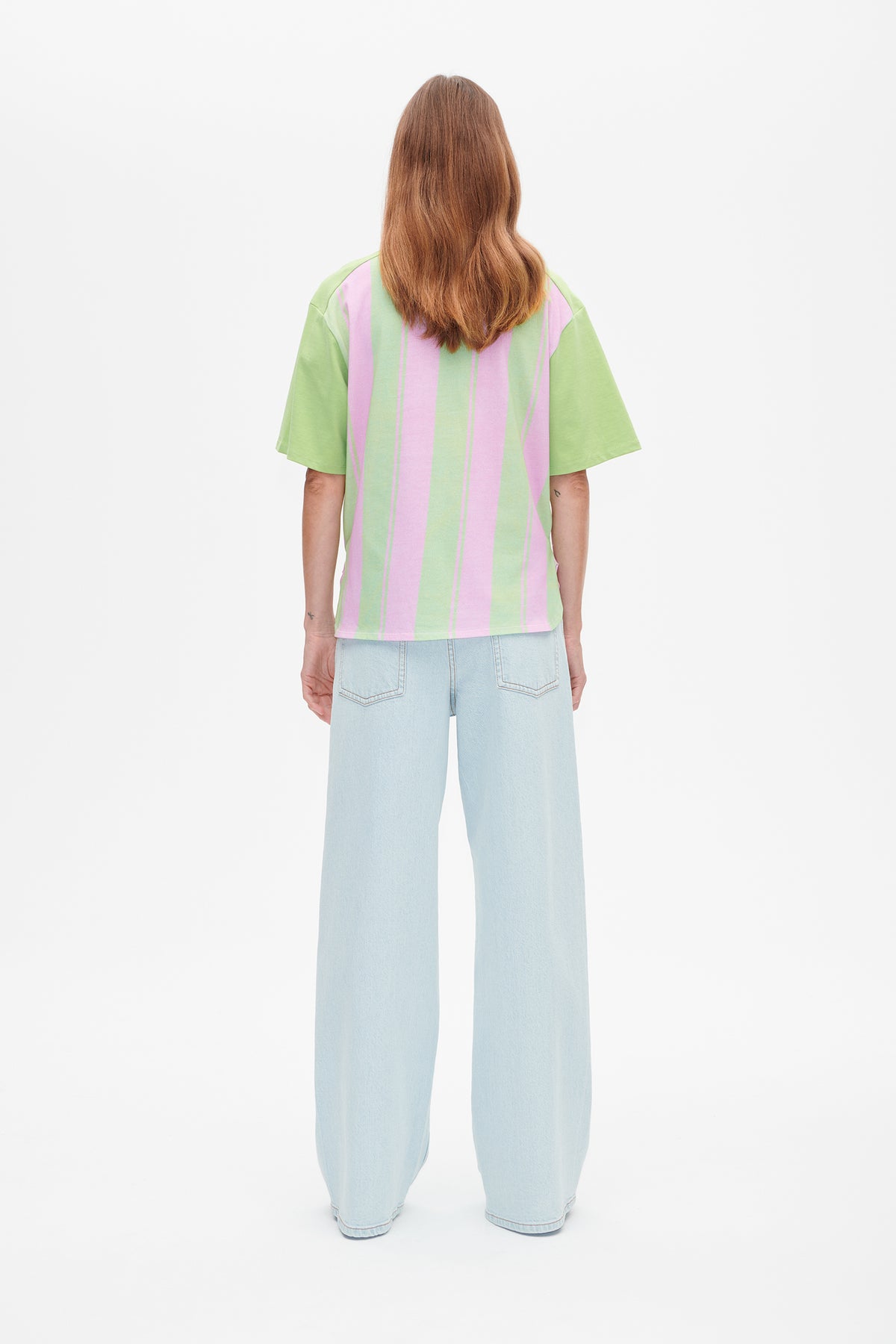 Vue de dos d'un T-shirt à manches courtes à larges rayures verticales rose et vert clair, porté avec un jean à coupe large bleu pâle.