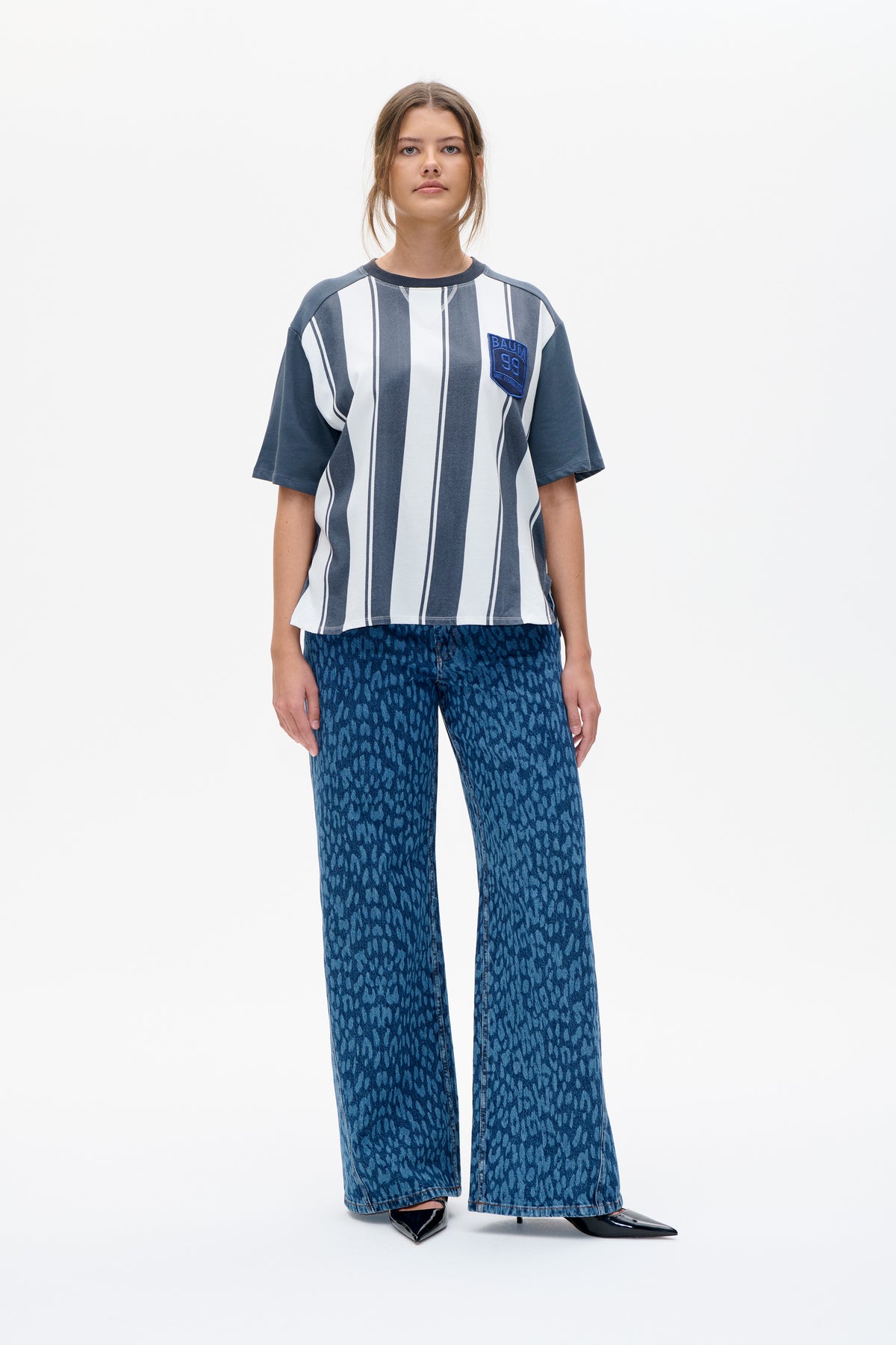 Front view of a model wearing a blue and white vertically striped short-sleeve T-shirt with a chest patch and patterned blue wide-leg jeans.
