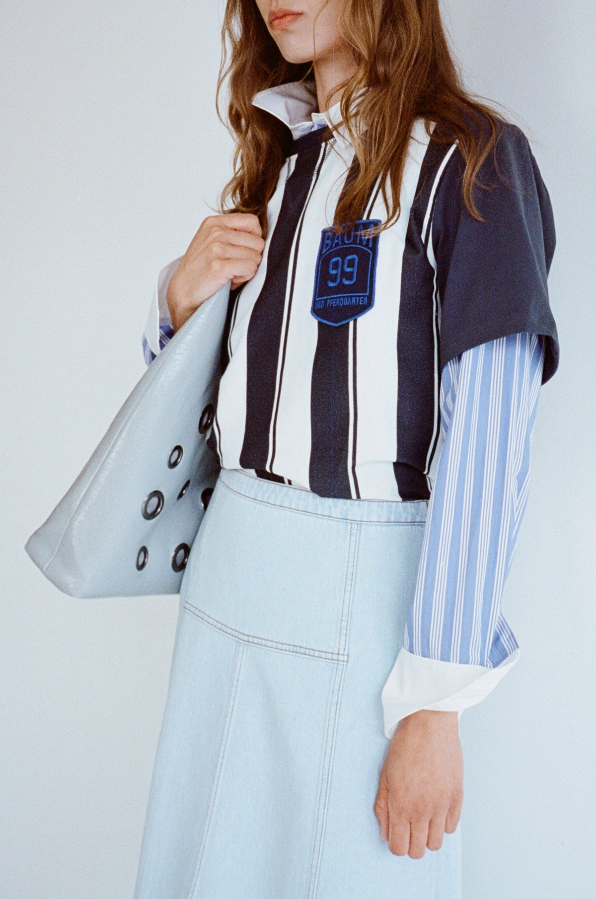 Cropped campaign view of a model wearing a navy-and-white striped sleeveless top over a blue striped shirt, with a pale denim skirt and gray tote bag.