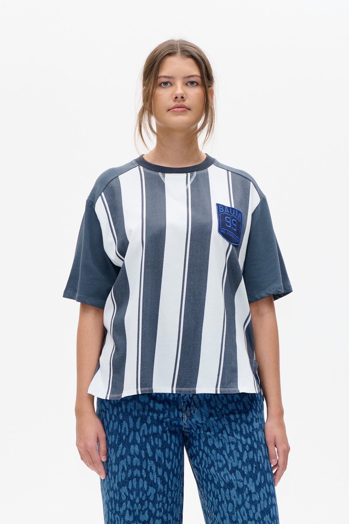 Front view of a model wearing a grey and white vertically striped short-sleeve top with a blue chest patch.