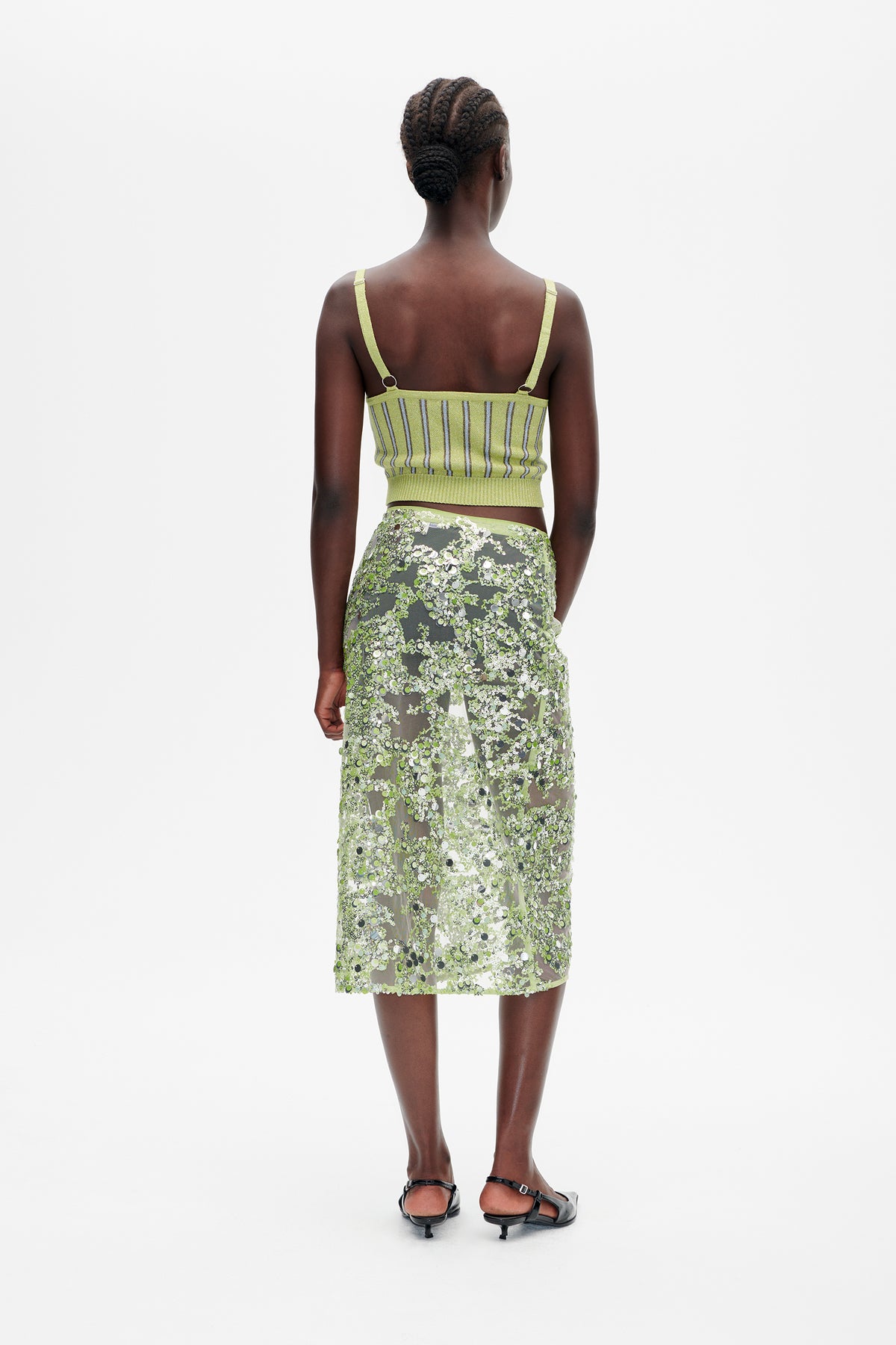 Rückansicht eines Models in einem limonengrünen, gestreiften Stricktop und einem transparenten, dicht mit Pailletten besetzten grünen Rock.