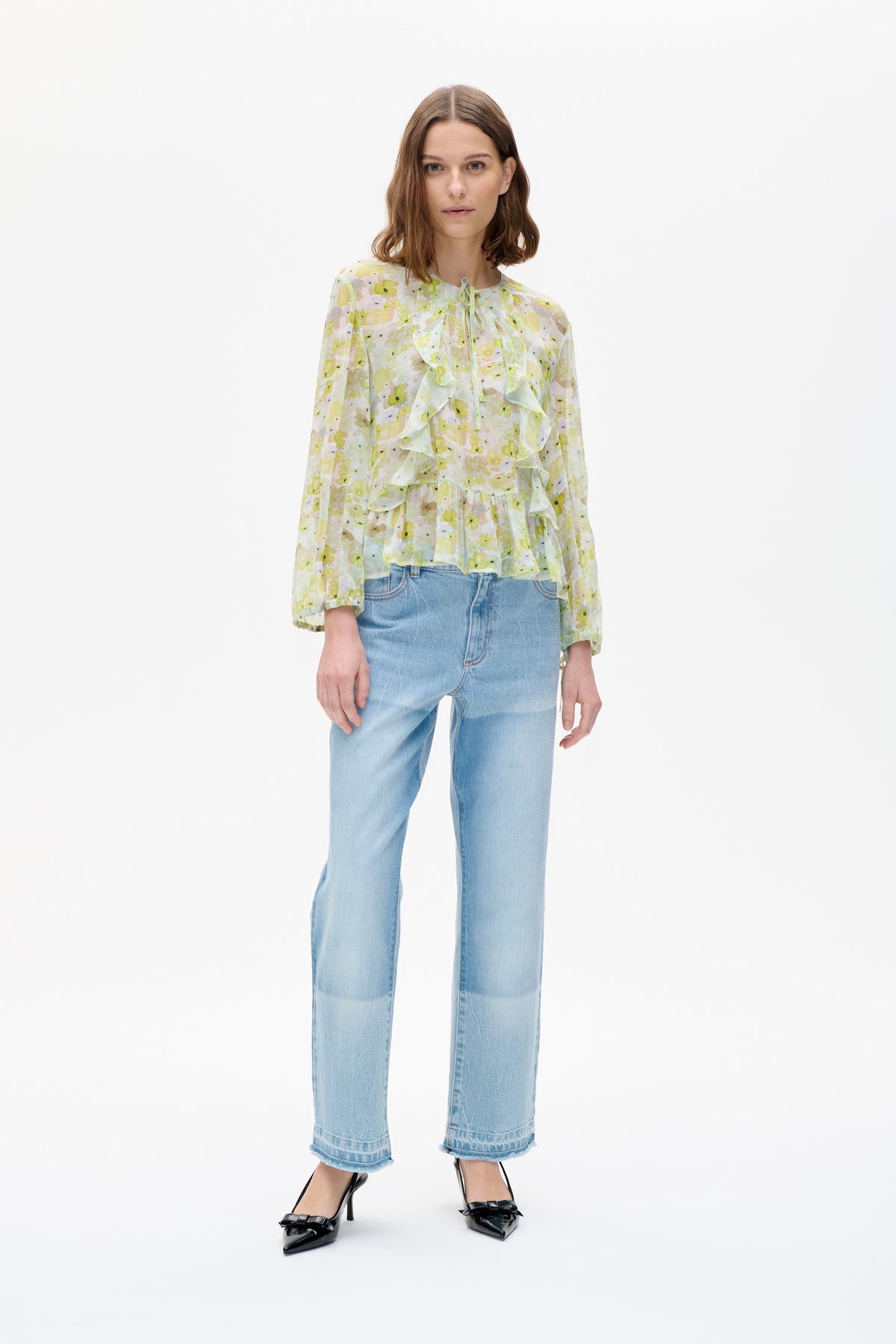 Front view of a pale yellow and green floral blouse with long sleeves, a tie neckline and cascading front ruffles, worn with light-wash jeans.