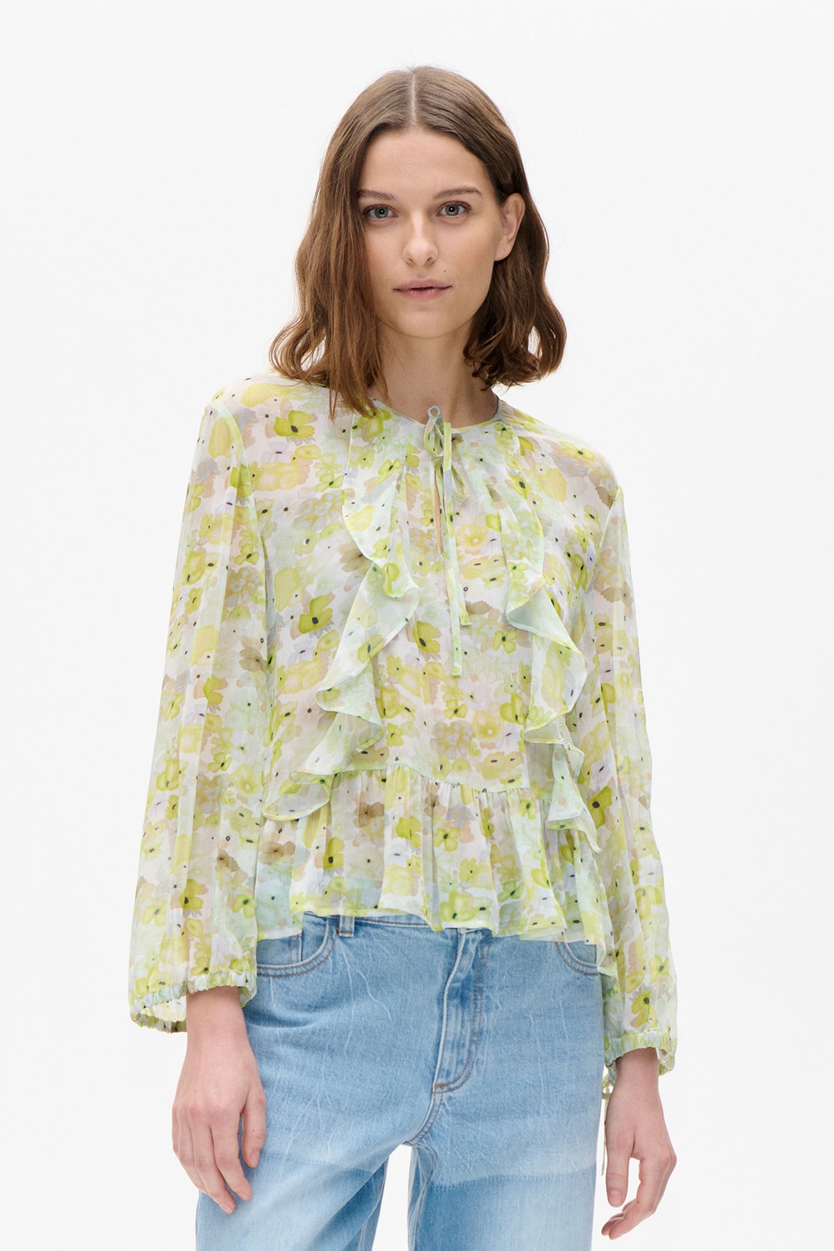 Vue de face d'une blouse pâle à imprimé floral abstrait jaune et vert, encolure à nouer, volants en cascade et manches trois-quarts froncées.