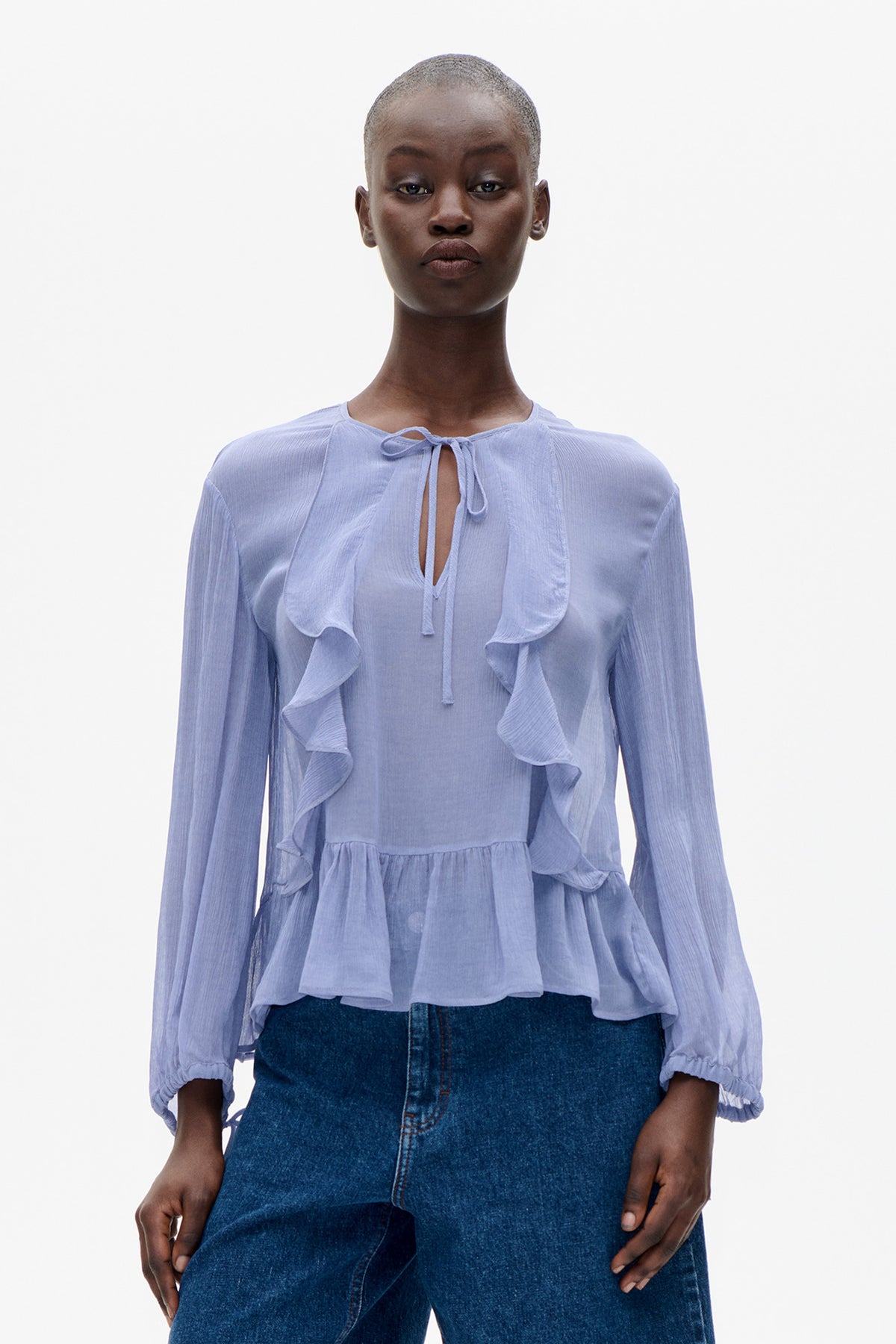 Vue de face d'une blouse bleu pâle à manches longues, encolure goutte à nouer, volants en cascade et poignets élastiqués, portée avec un jean foncé.