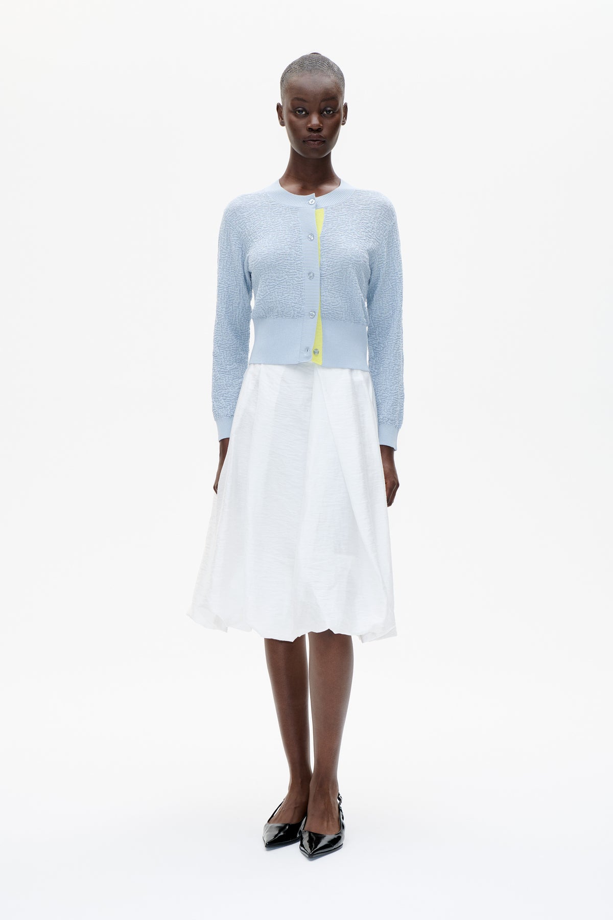 Frontalansicht eines Models, das eine hellblaue, strukturierte Strickjacke mit Knopfleiste über einem weißen Kleid trägt, gestylt mit schwarzen spitzen Schuhen.