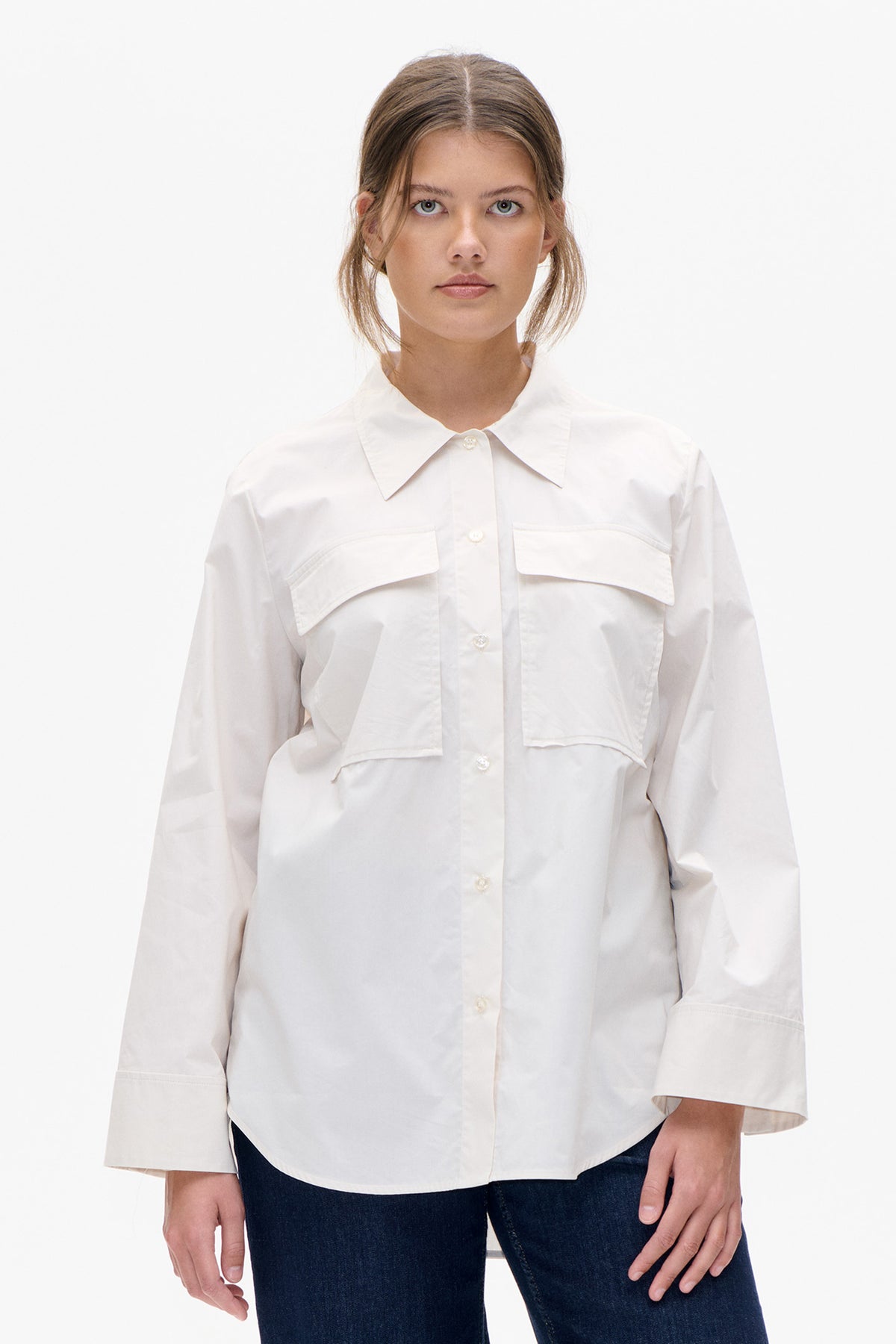 Vue de face d'une chemise blanche boutonnée sur le devant, col pointu oversized, manches courtes et deux poches poitrine à rabat angulaires, portée avec un jean foncé.