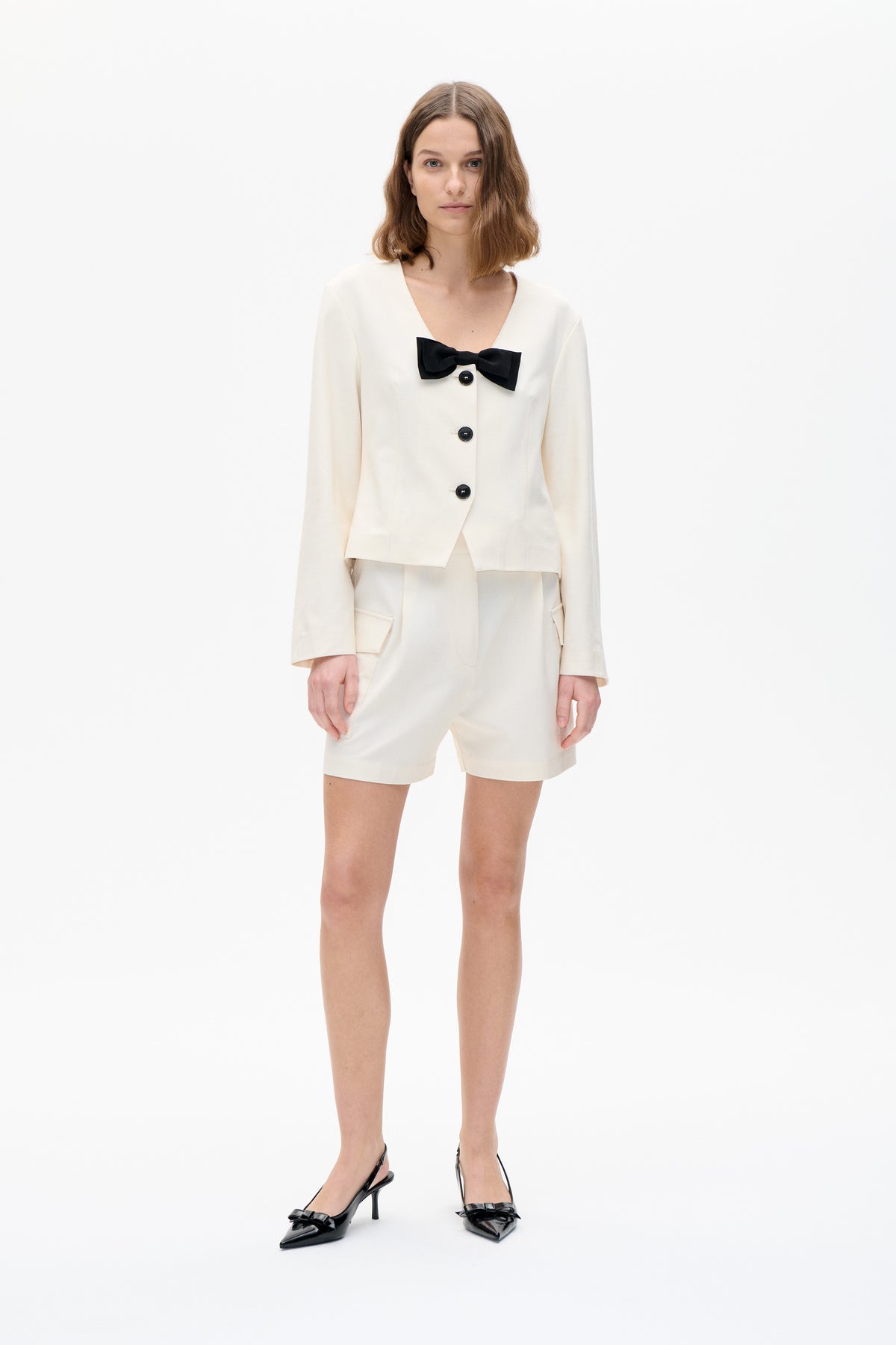 Model wearing an ivory jacket with a black bow and buttons, paired with matching shorts; front view against a white background.