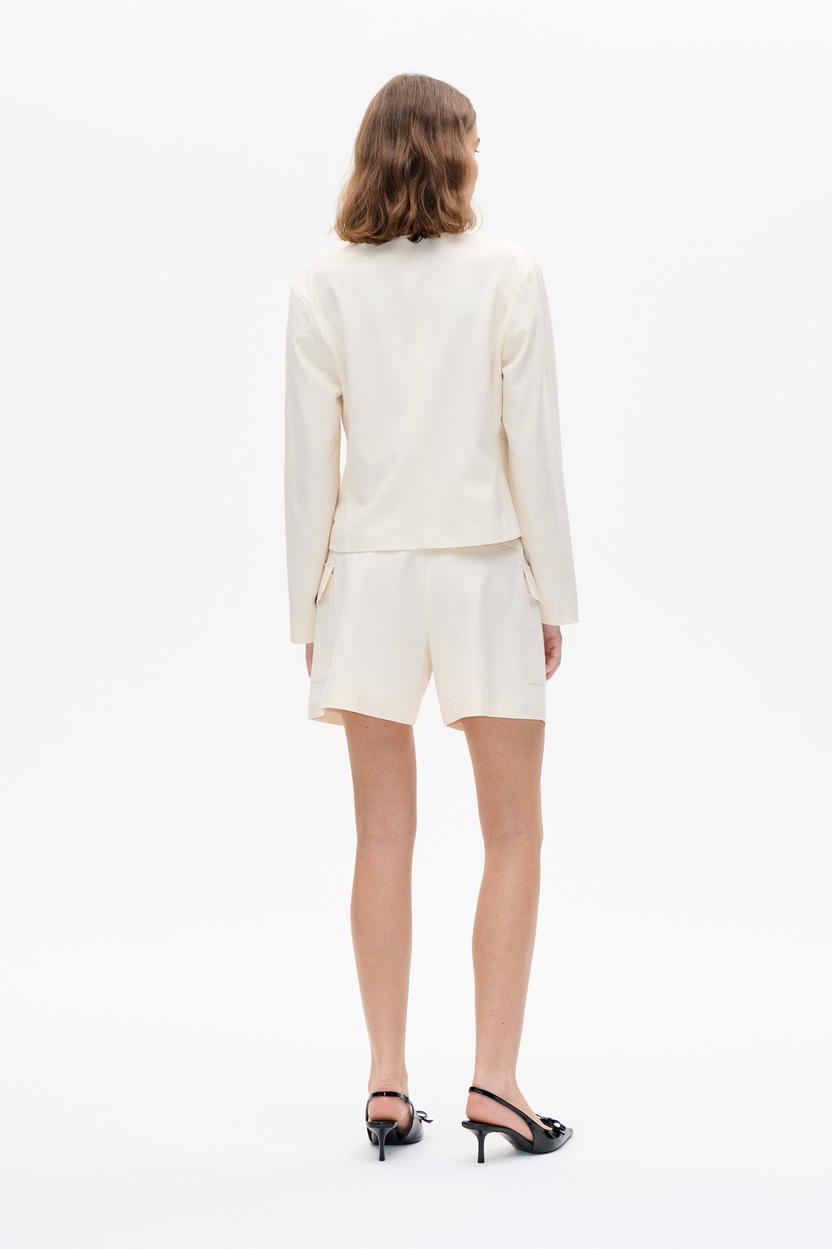 Back view of a model in a cream long-sleeved jacket and matching tailored shorts with side pockets, paired with black slingback heels.