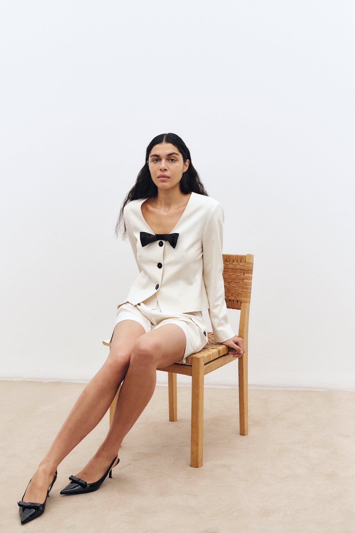 A seated model wears an ivory button-front jacket with a black bow detail, matching shorts and black bow flats against a pale backdrop.