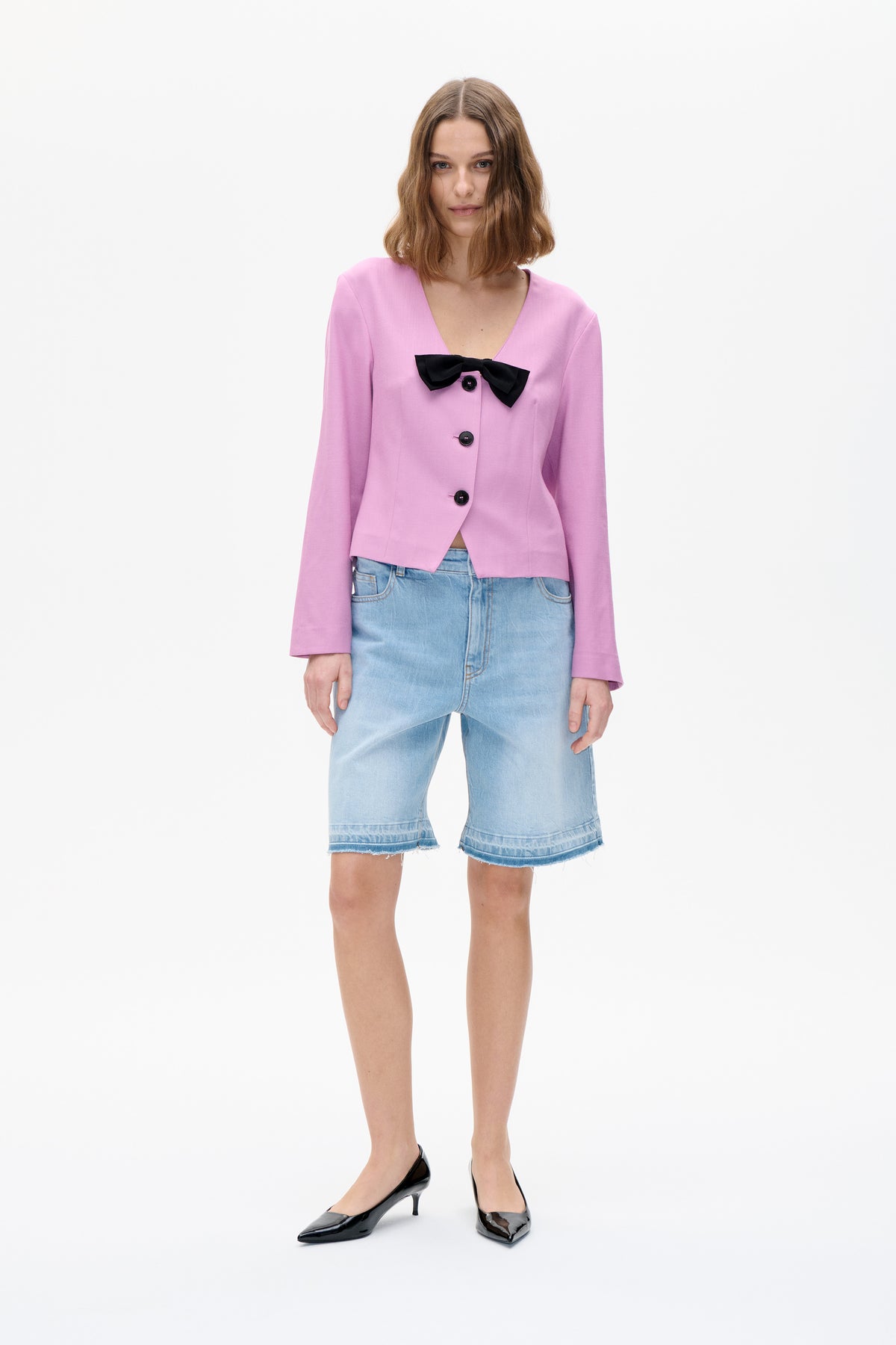 Front view of a model wearing a pink V-neck jacket with black bow and buttons, paired with light-wash denim shorts.