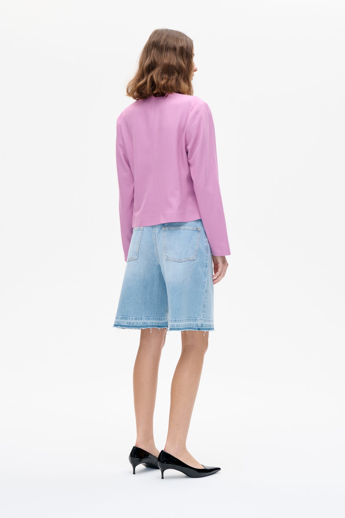 Back view of a model wearing a plain lilac long-sleeved top with light-wash denim shorts and black pointed heels.