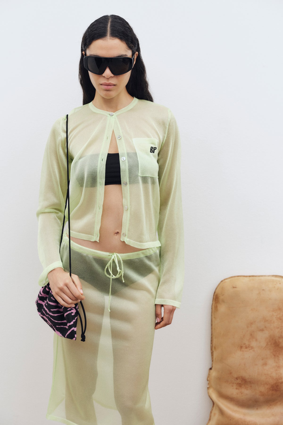 Campaign portrait of a model in a sheer pale lime button-front cardigan and matching drawstring trousers, with a black crop top and oversized sunglasses.