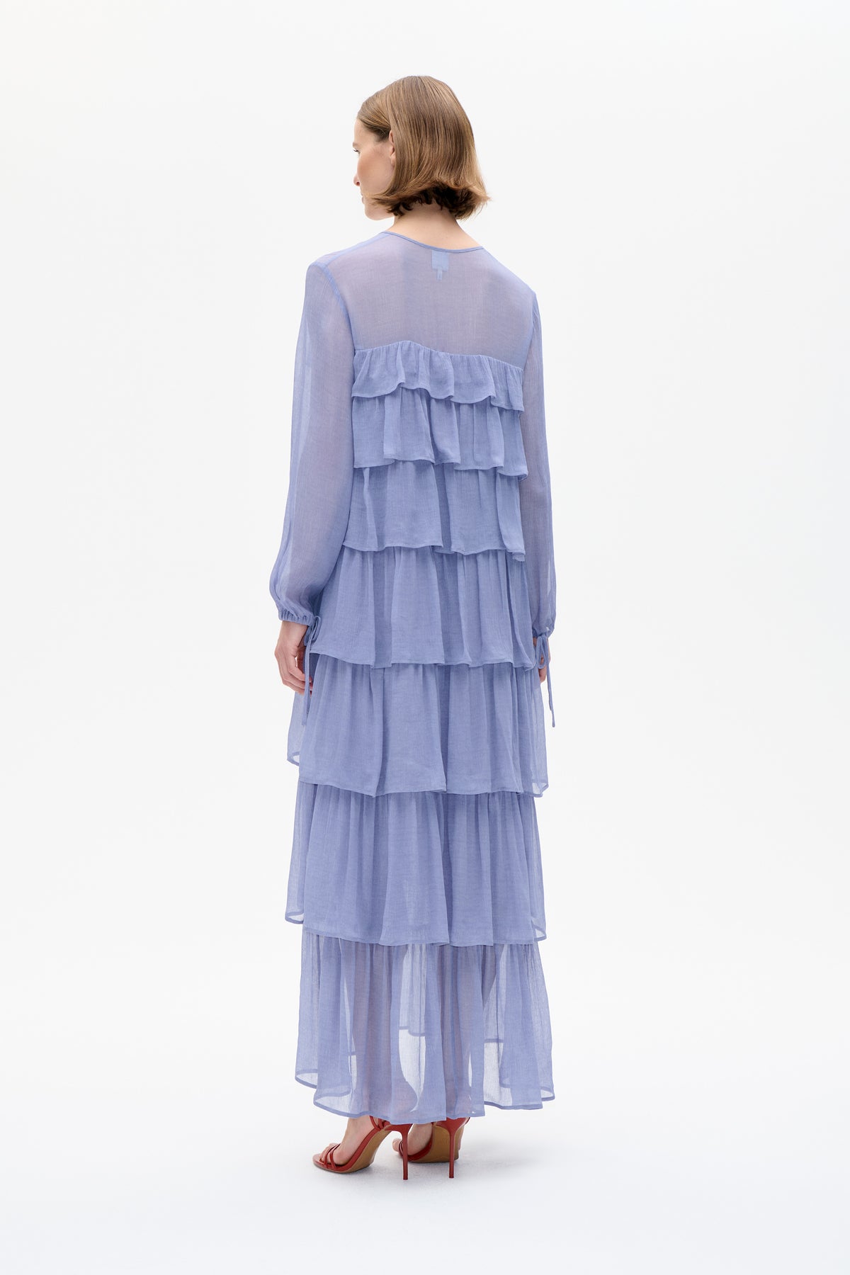 Back view of a model wearing a sheer lavender long-sleeved dress with layered ruffles and a flowing tiered skirt.