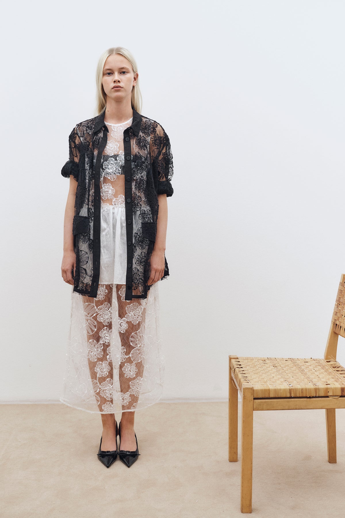 Le mannequin se tient à côté d'une chaise tressée, portant une chemise noire transparente en dentelle florale, boutonnée, par-dessus une robe blanche en dentelle florale.