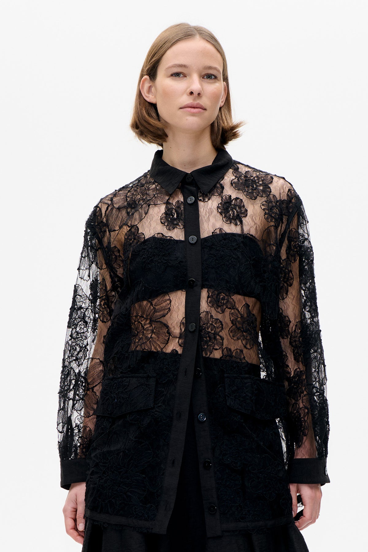 Détail avant d'une blouse noire boutonnée sur le devant, avec manches et empiècements en dentelle florale transparente, bandes noires opaques, col pointu et larges poignets.