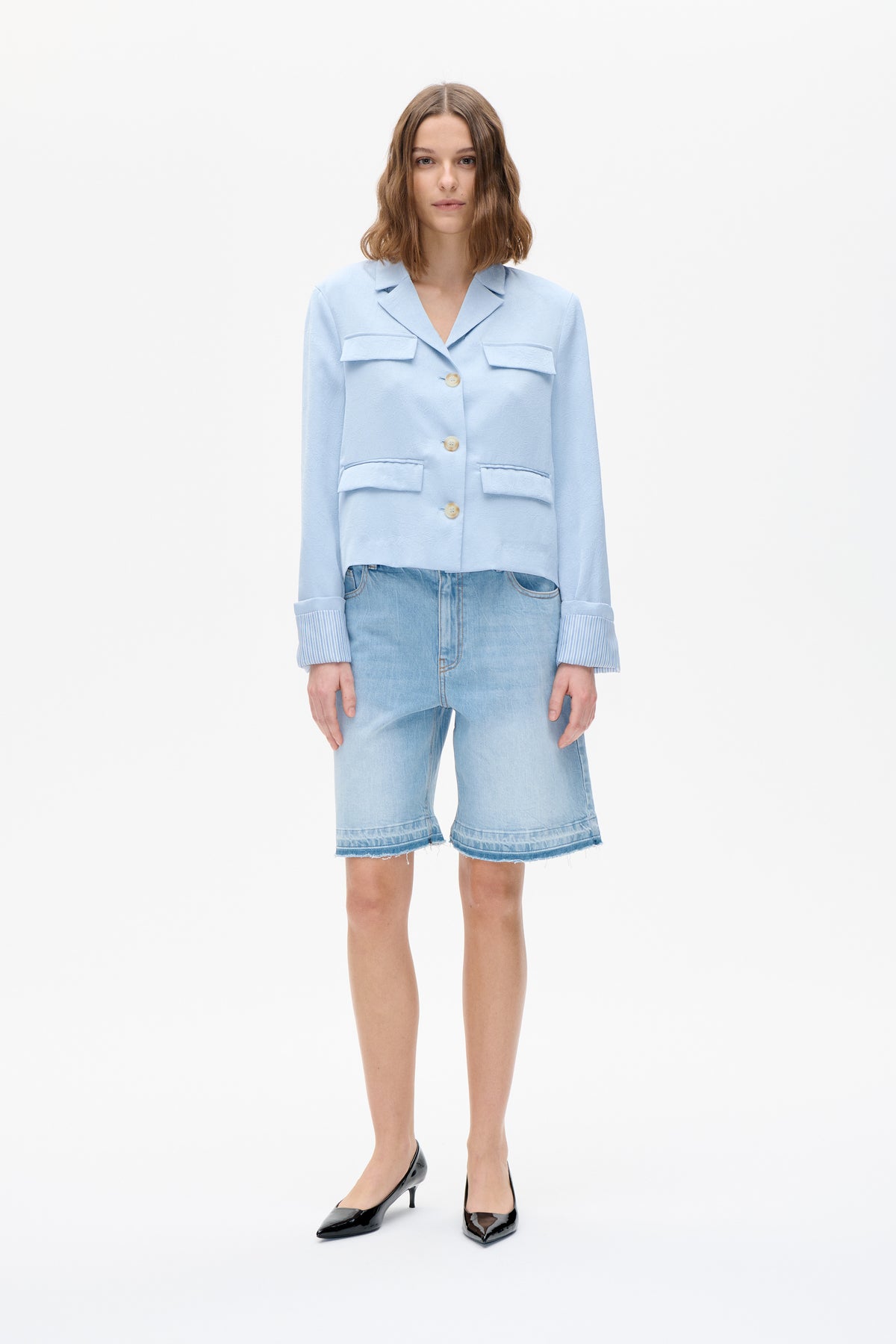Front view of a model wearing a pale blue textured button-front jacket with four flap pockets and striped cuffs, paired with denim shorts.