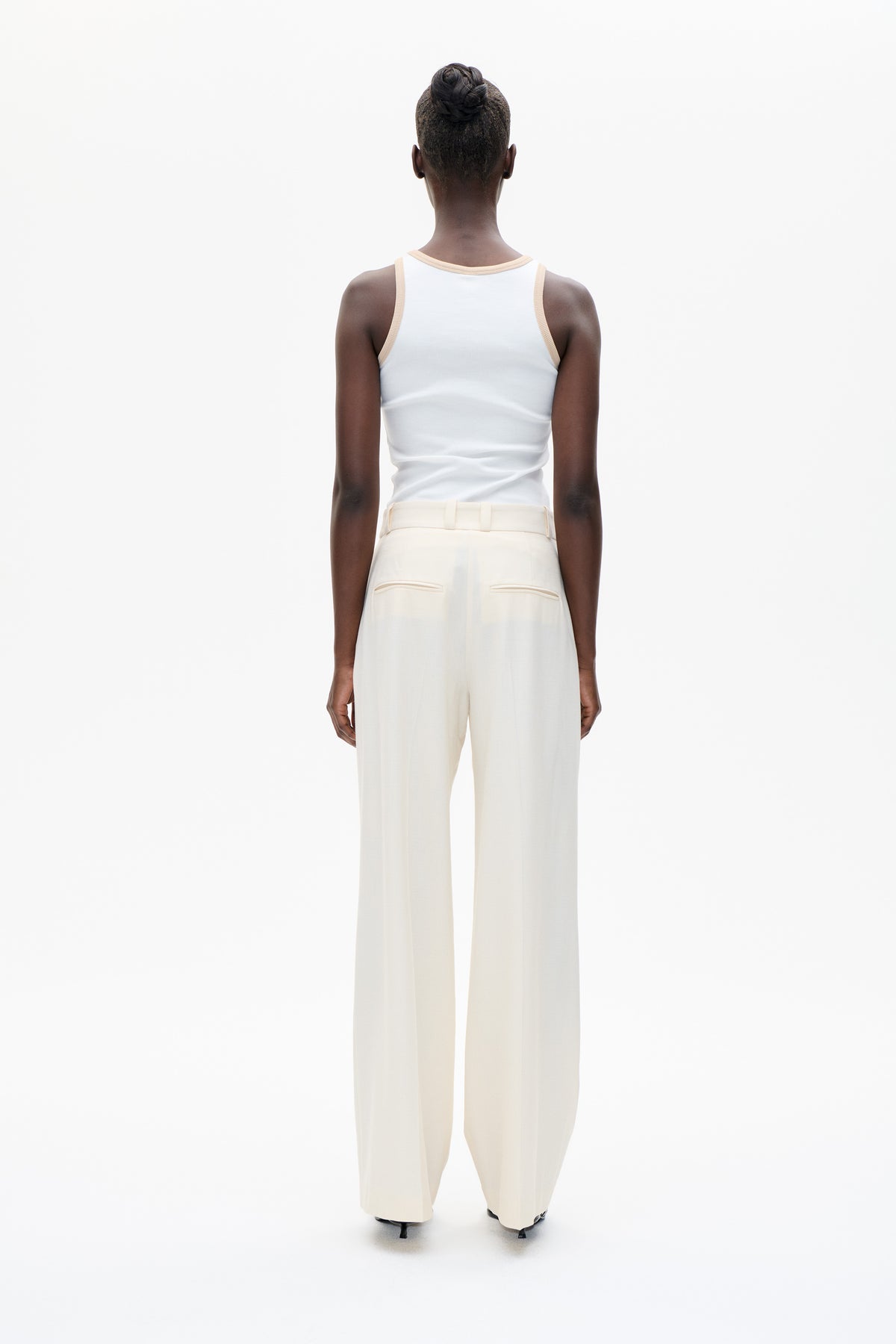 Rear view of a model in a white tank top with pale beige edging and cream wide-leg trousers with piped back pockets.