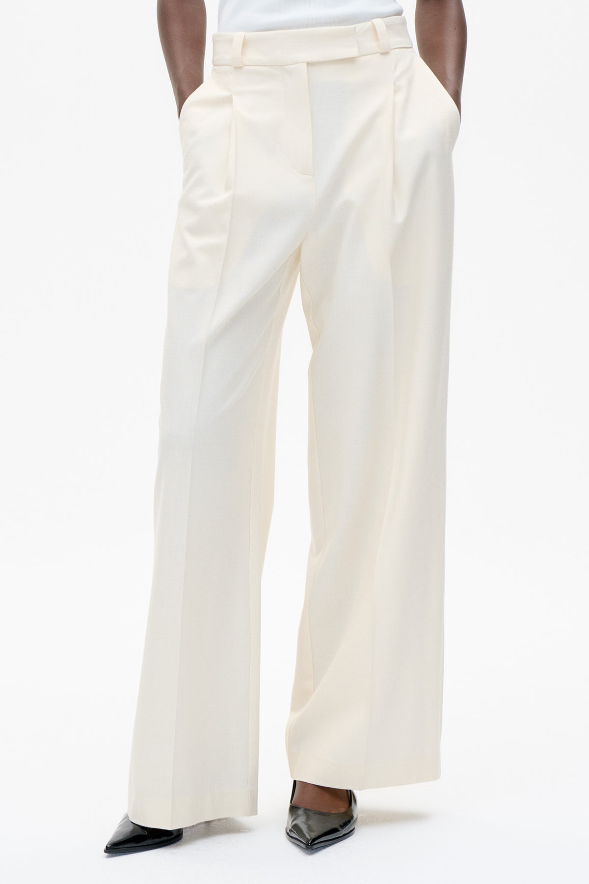 Front detail of cream wide-leg trousers with a high waistband, belt loops and front pockets, styled with black pointed shoes.