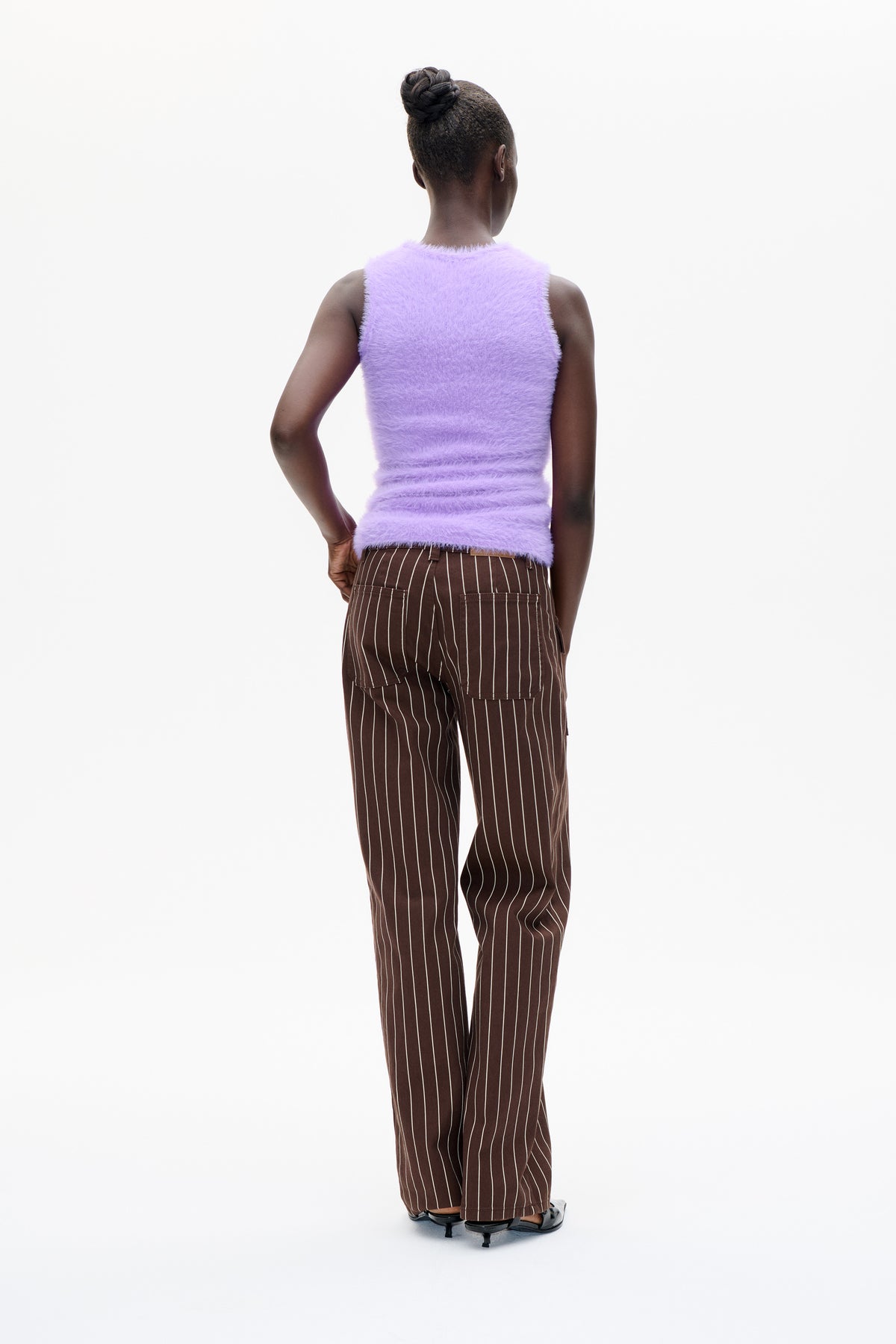 Achteraanzicht van een model in een lila, pluizige mouwloze top en een bruine broek met dunne witte verticale strepen en opgestikte zakken.