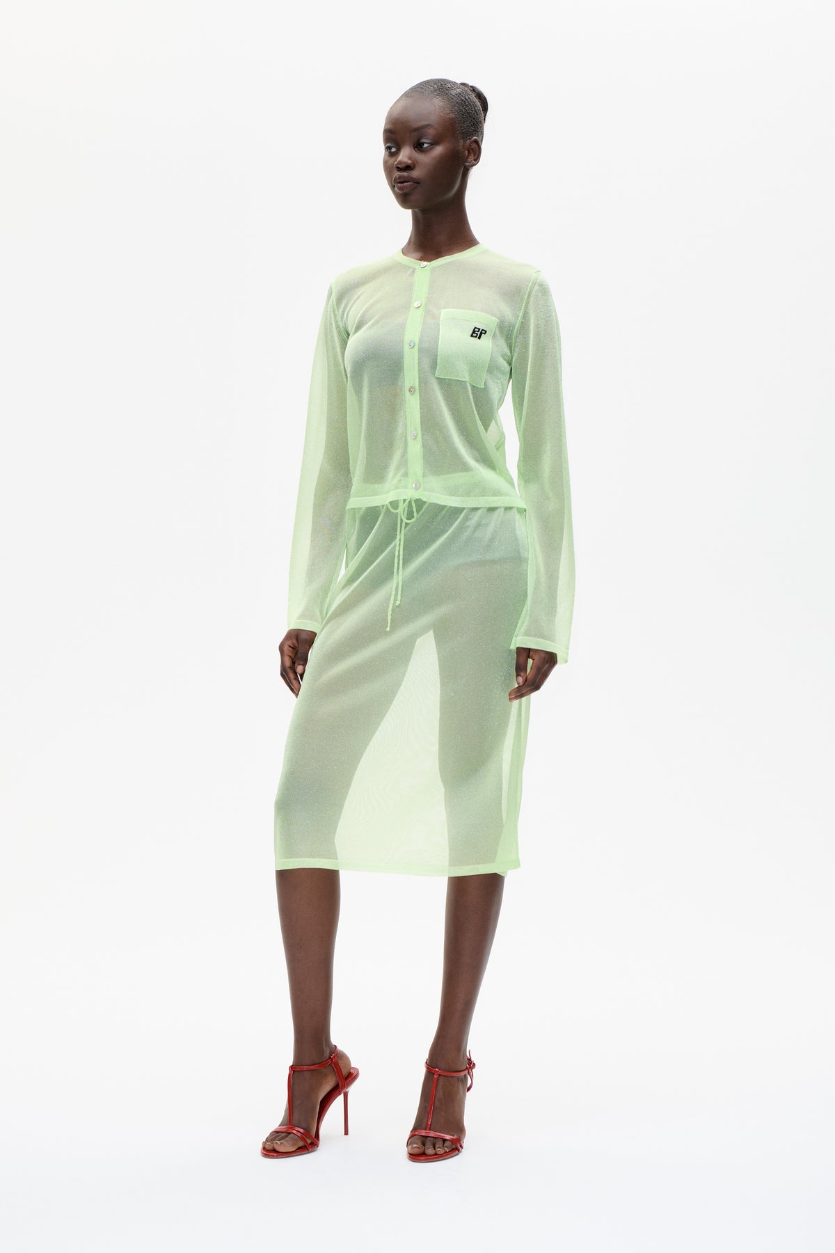 Model wearing a sheer pale-green button-front cardigan with chest pocket and matching sheer skirt, styled with red strappy heels.