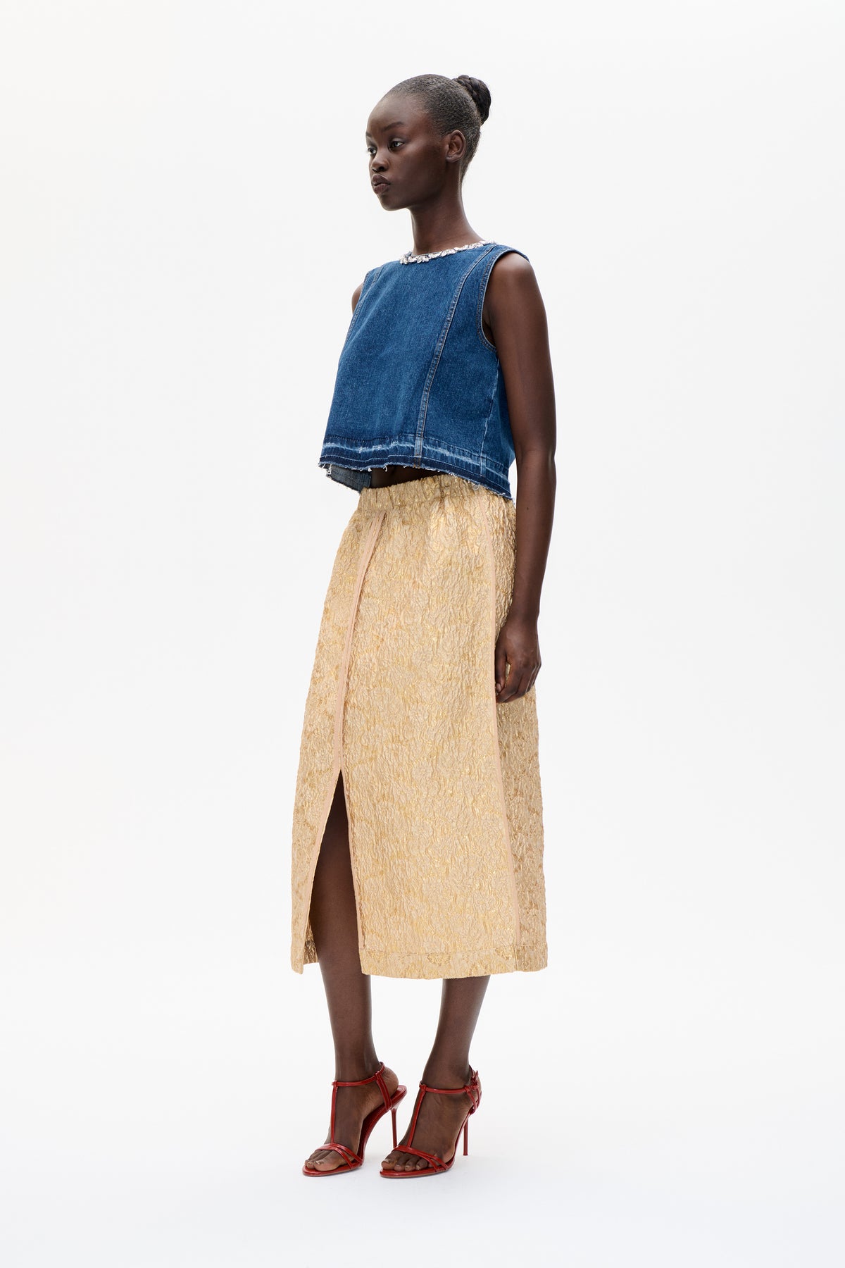 Model draagt een blauwe mouwloze denimtop met versierde halslijn en onafgewerkte zoom, een gouden midi-rok met structuur en rode pumps tegen een witte achtergrond.
