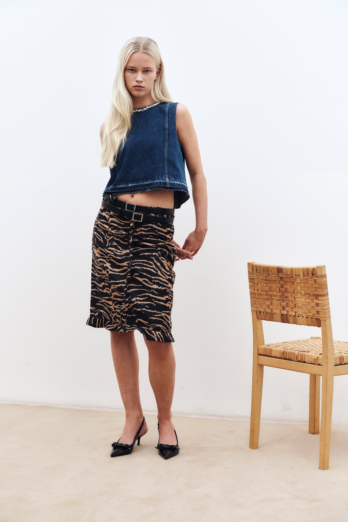 Campagneportret van een model in een donkerblauwe mouwloze denimtop met een halslijn met sierstenen en een rok met zwart-bruin patroon naast een gevlochten stoel.