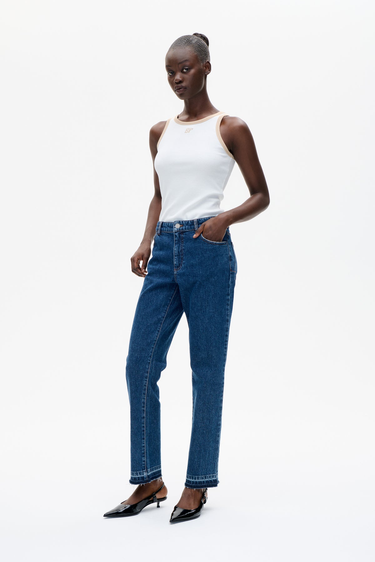 Model trägt ein weißes ärmelloses Top mit beigen Besätzen und blaue Straight-Leg-Jeans mit ausgefransten Säumen; dazu schwarze High-Heels.