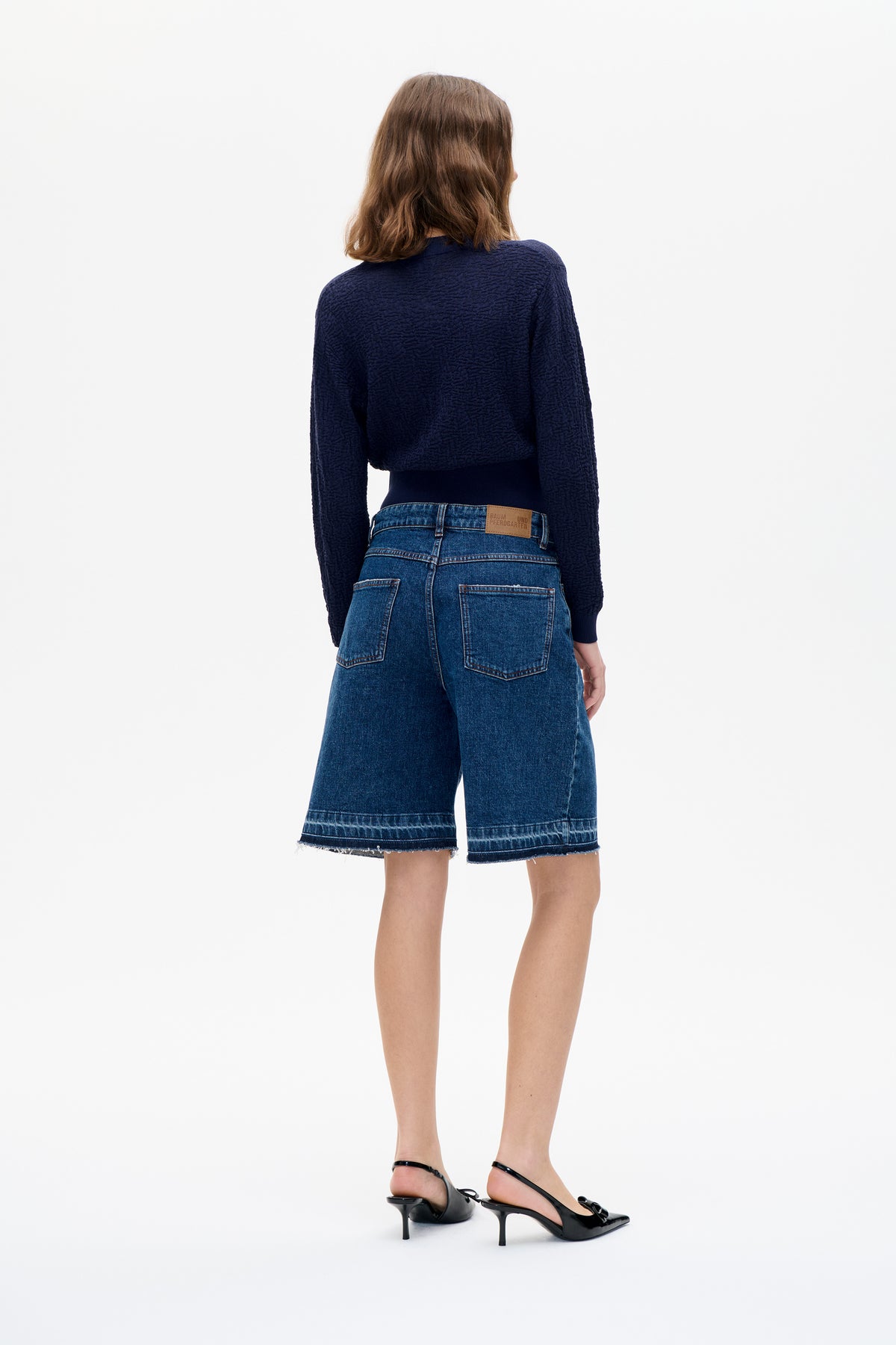 Rear view of a model wearing a navy textured long-sleeve sweater and blue denim knee-length shorts with raw hems.