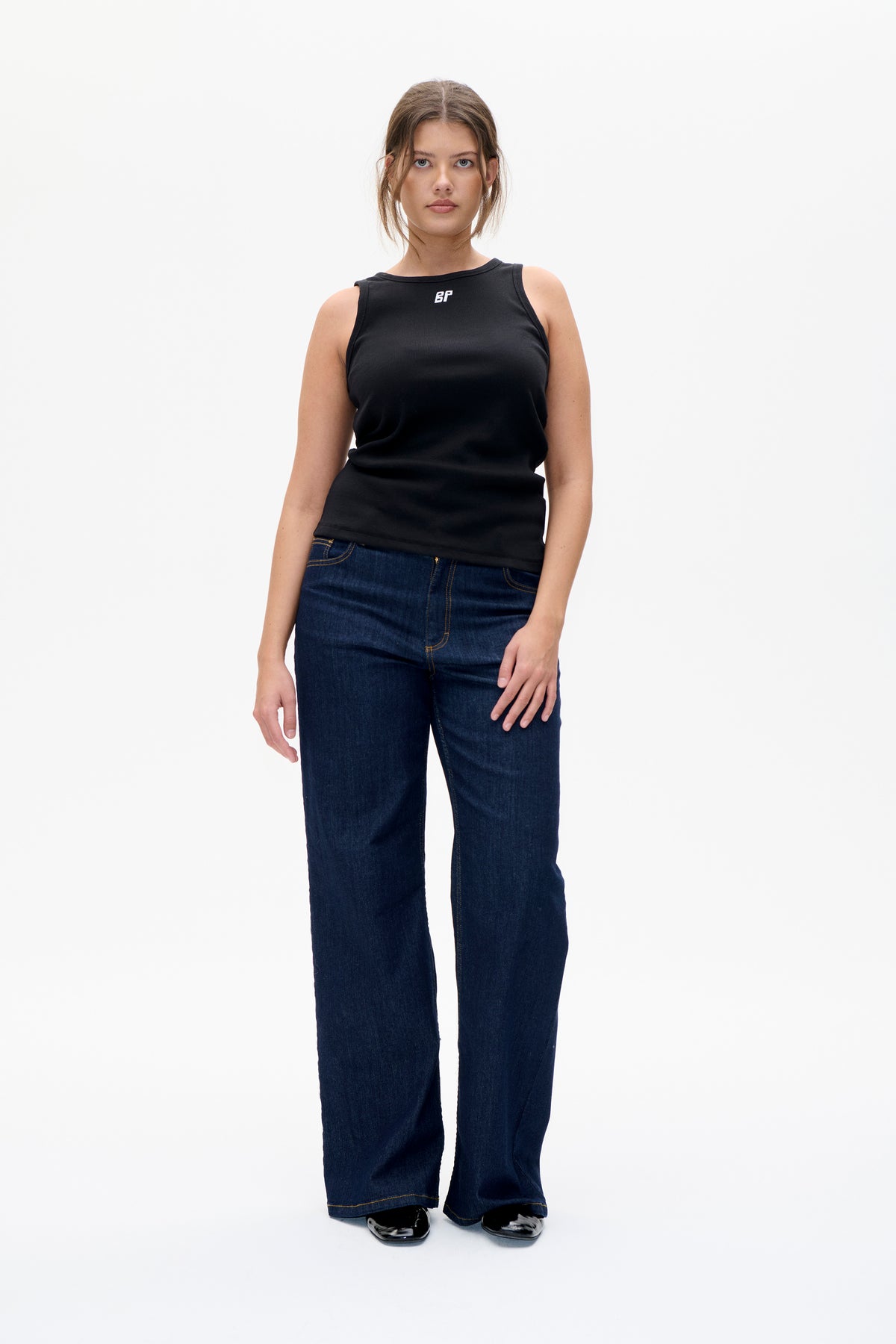Vooraanzicht van een model in een zwarte mouwloze top met een klein wit borstlogo en donkerblauwe wijdvallende jeans.