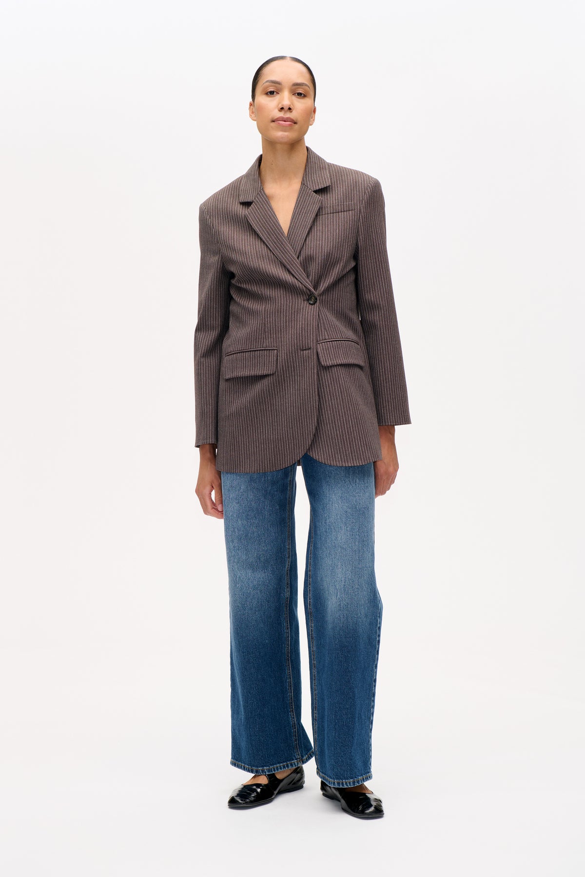 Model draagt een bruine blazer met fijne verticale strepen, ingesneden revers en klepzakken, gecombineerd met blauwe jeans met wijde pijpen.