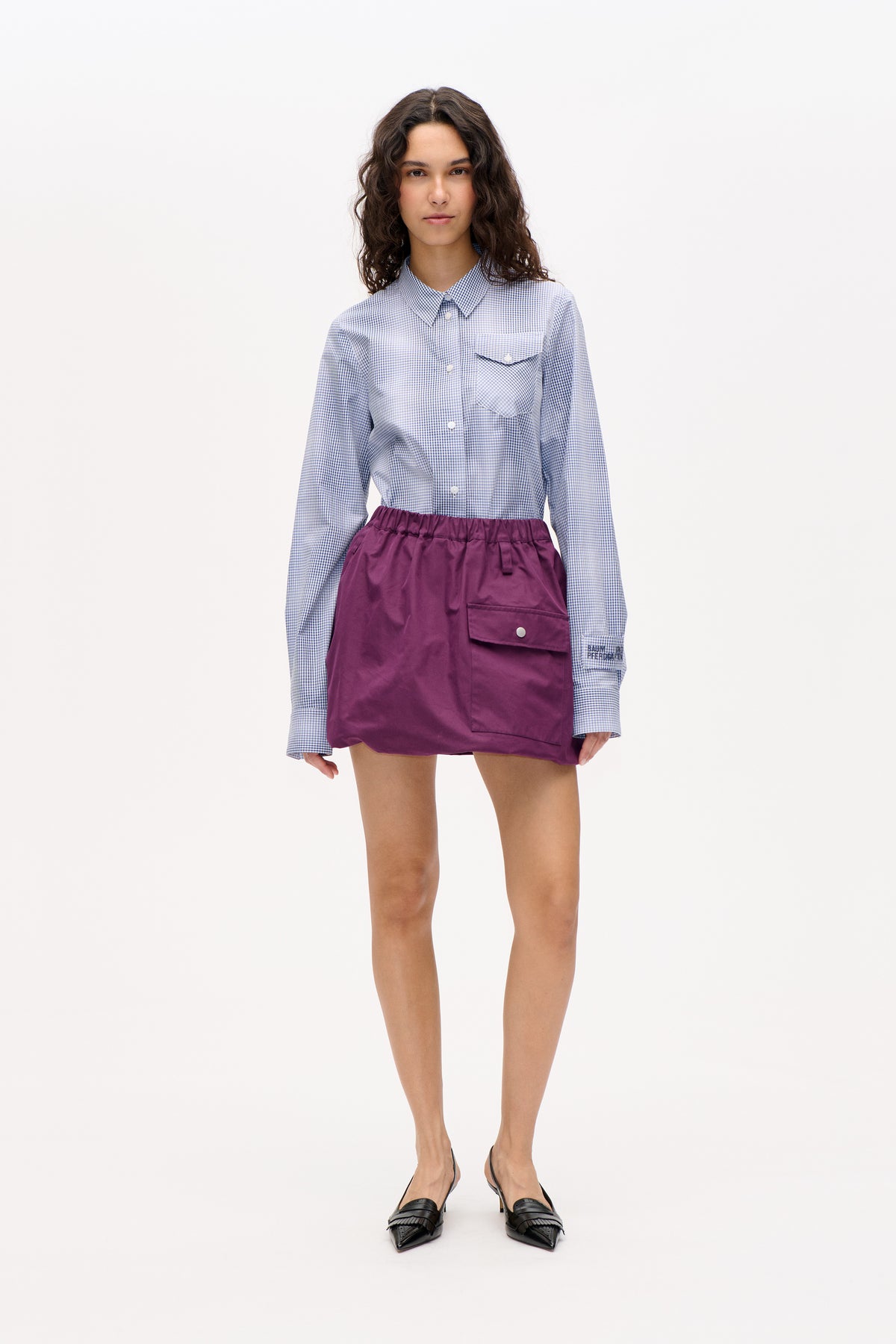 Vorderansicht eines Models in blau-weißem Vichy-Hemd mit Knopfleiste und Brusttasche sowie pflaumenfarbenem Minirock mit großer Klappentasche.
